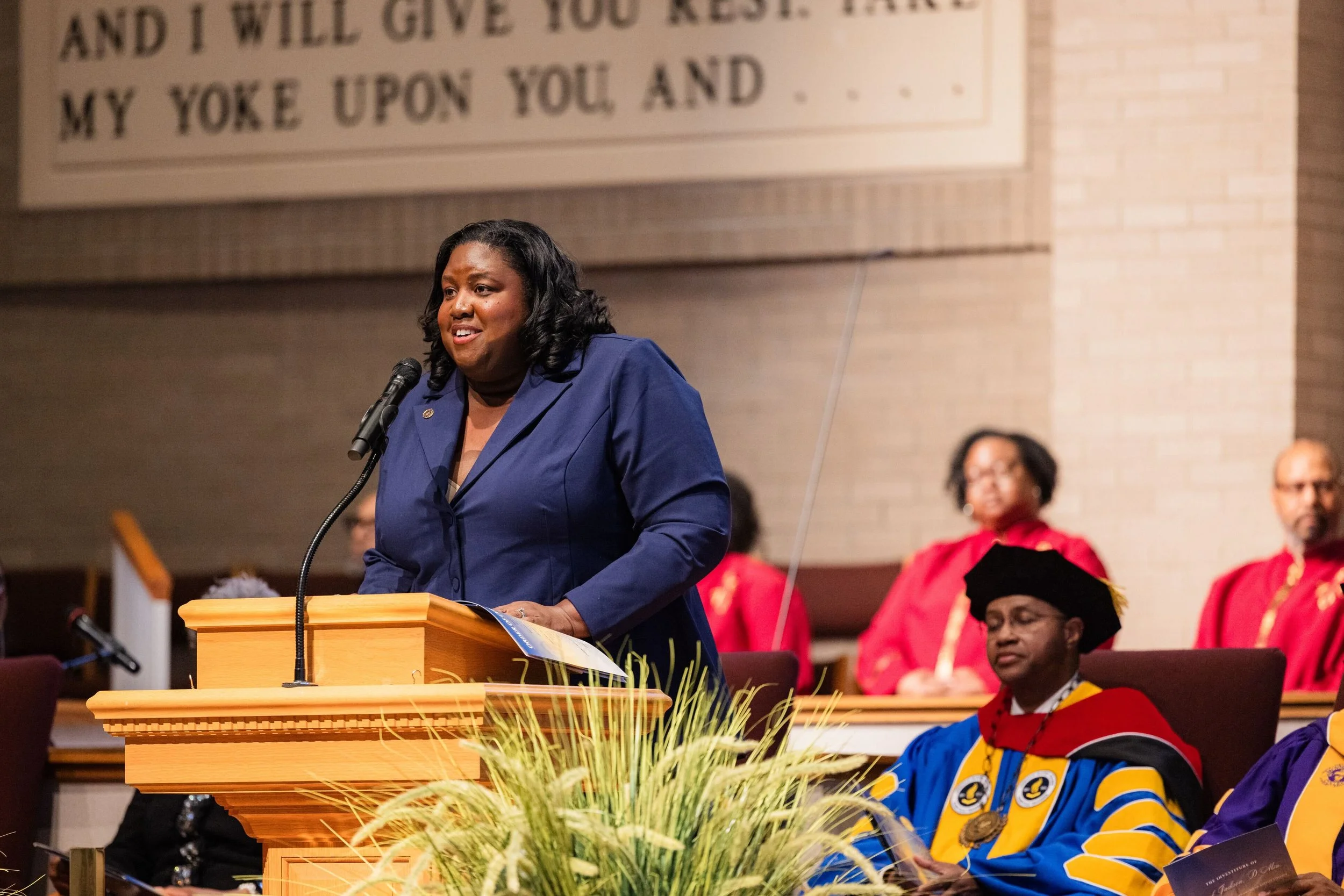 Monchiere' Holmes Jones, Cultural Economist™ speaking at a ceremony at American Baptist College with African American individuals in academic regalia sitting behind her. A large banner in the background and some people are wearing red robes.