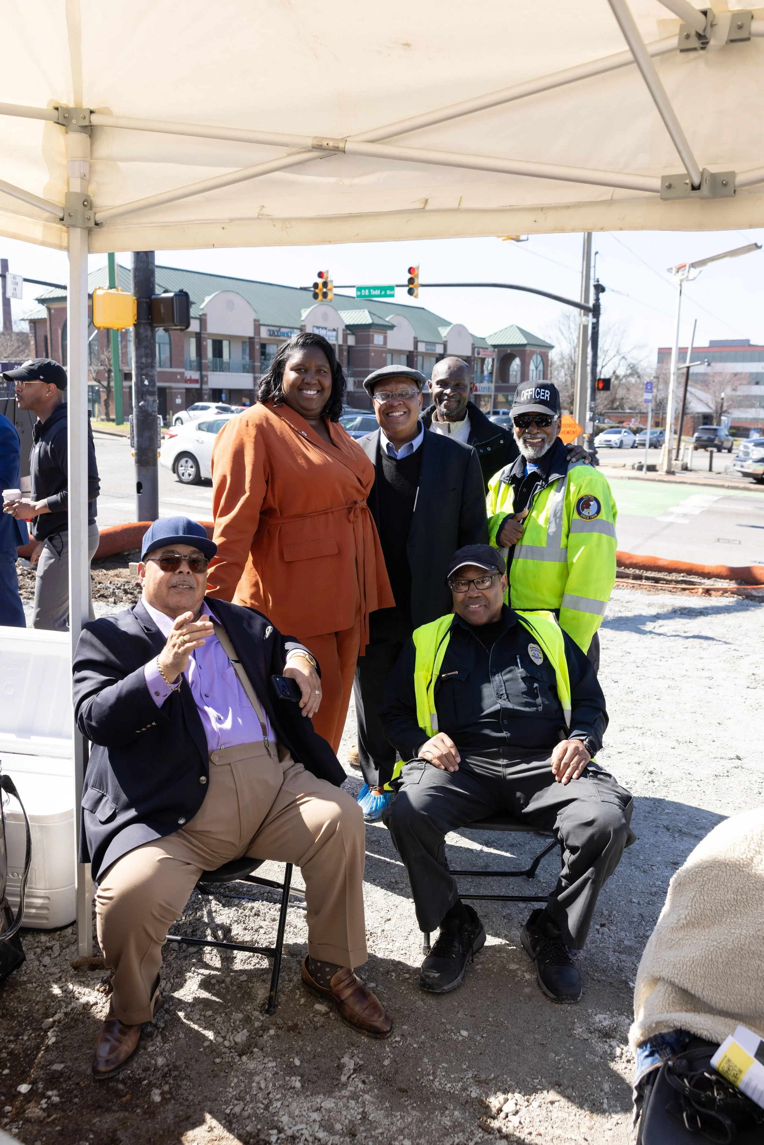 Monchiere' Holmes Jones, Cultural Economist™  with a  group of smiling men and women, gathered outdoors under a white canopy tent, on a city street with commercial buildings, traffic lights, and parked cars in the background.