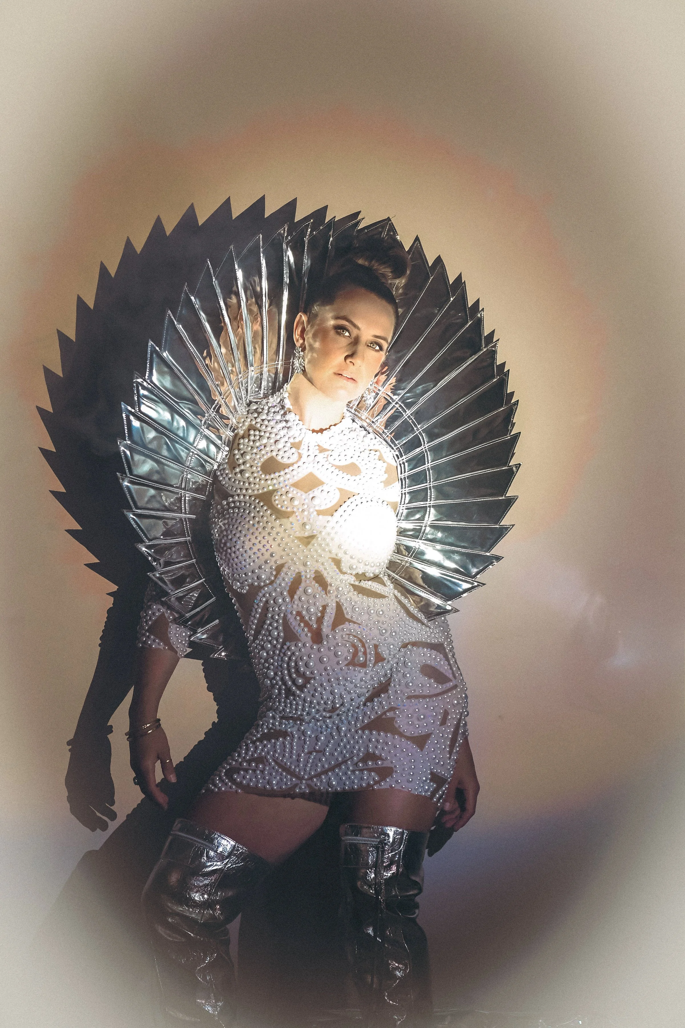Woman wearing an ornate, pearl-adorned dress with metallic thigh-high boots, posing with a large, reflective, fan-like accessory.
