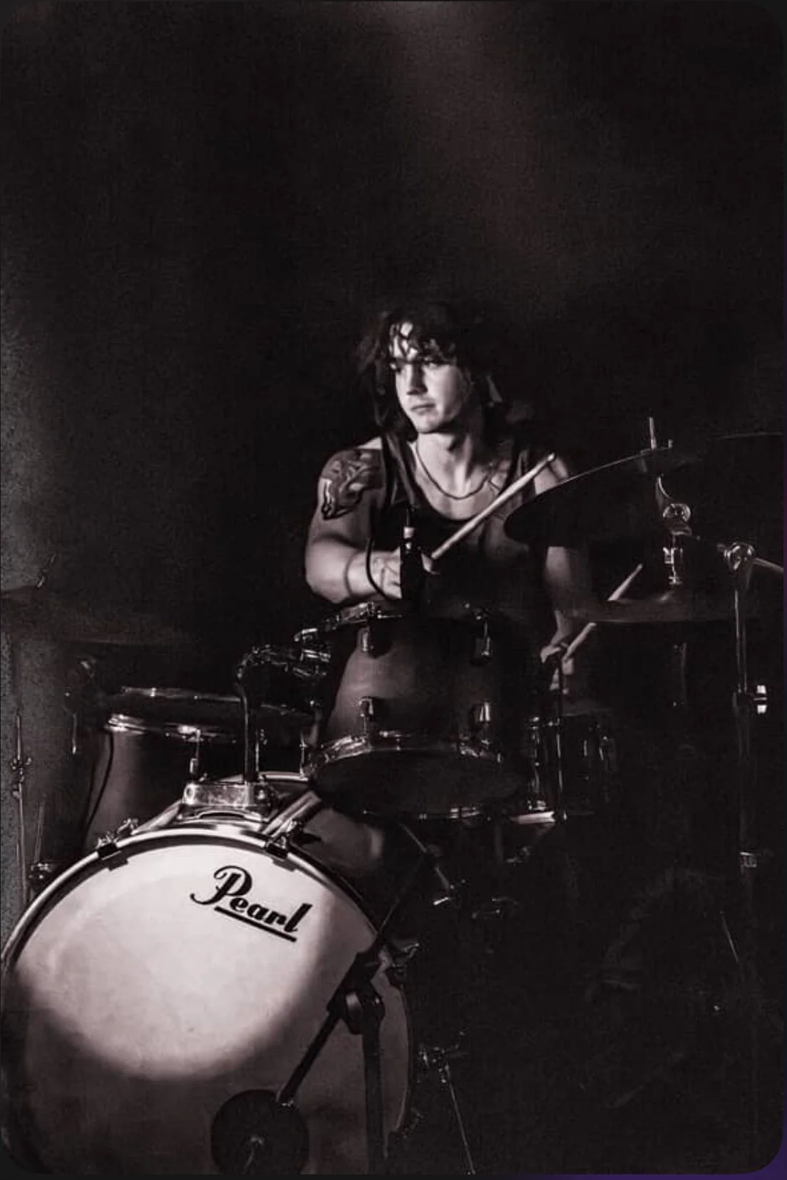 A person playing a drum set in a dark room, with a focused expression, wearing a sleeveless shirt, and tattoos on their arms.