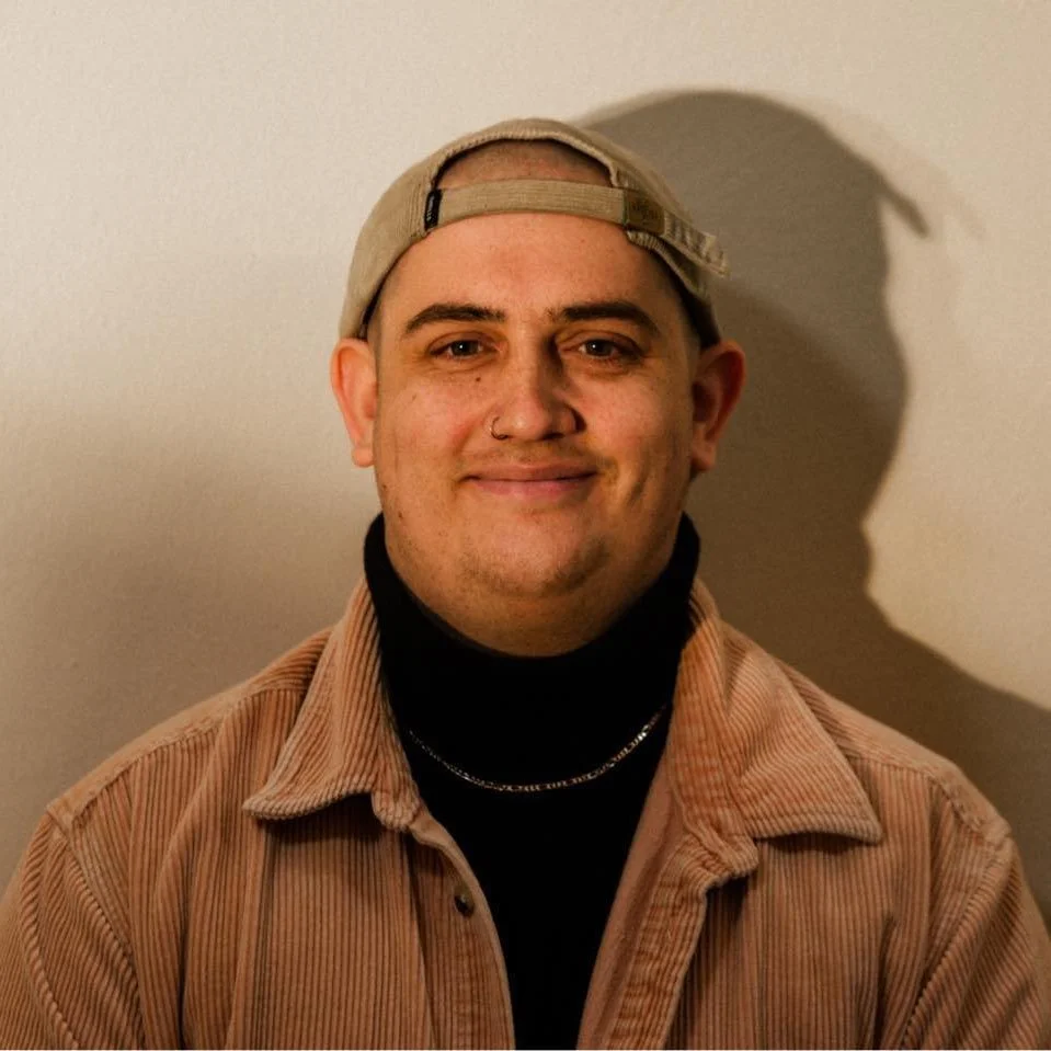 A young man wearing a beige baseball cap backward, black turtleneck, brown corduroy jacket, and a silver chain, smiling at the camera with a shadow cast on a plain wall behind him.