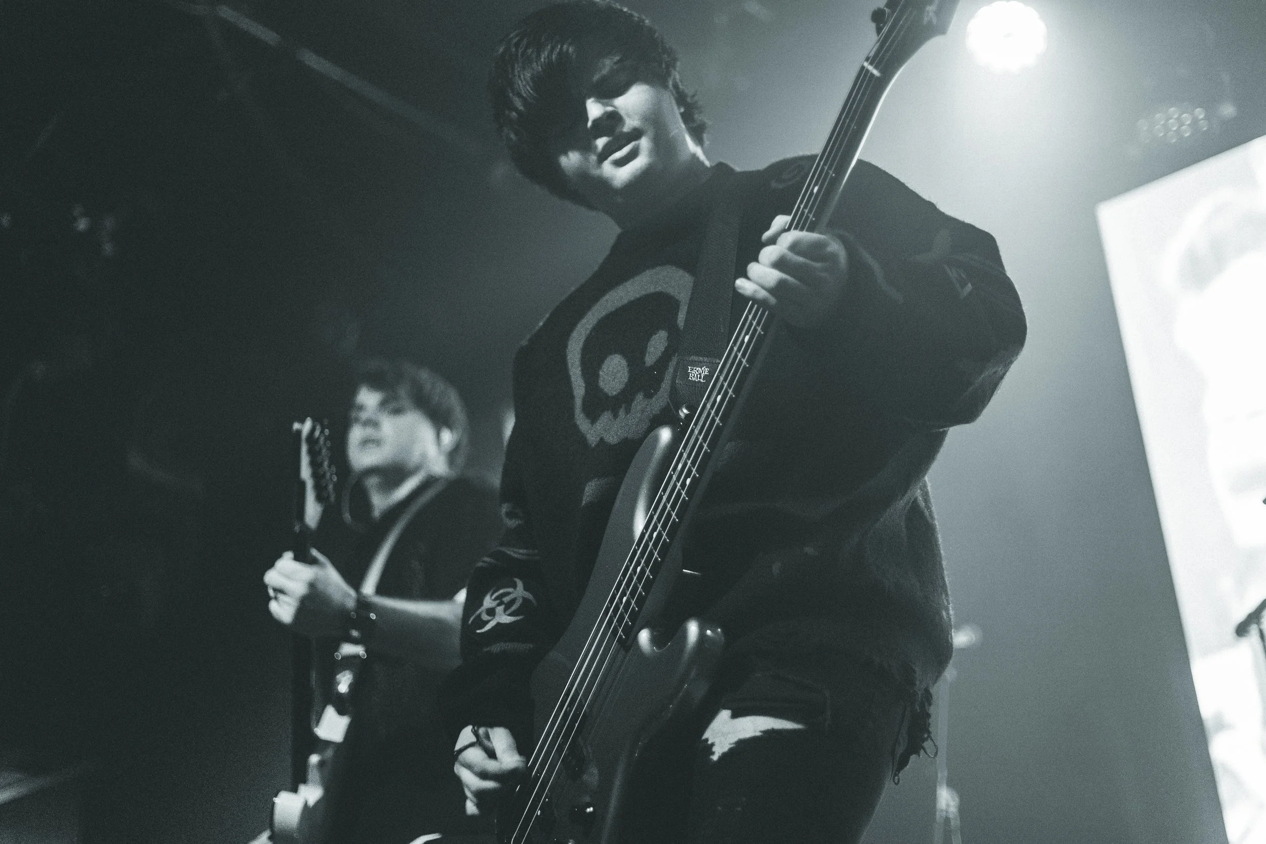A black-and-white photo of a musician playing an electric guitar on stage, with another person in the background also holding a guitar.