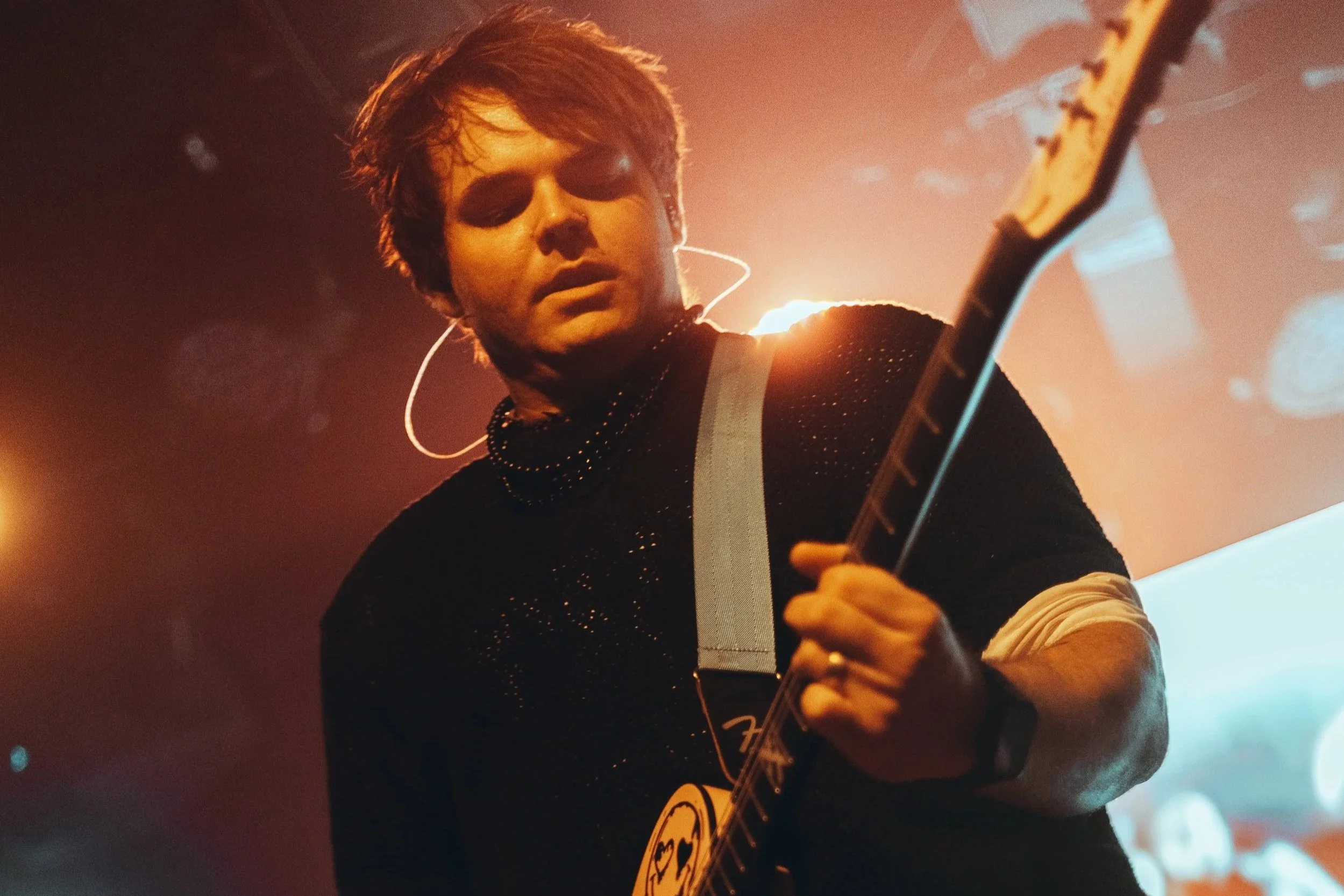 A young person with short hair wearing earrings and a black sparkly top, playing an electric guitar on stage with warm lighting and a blurry background.
