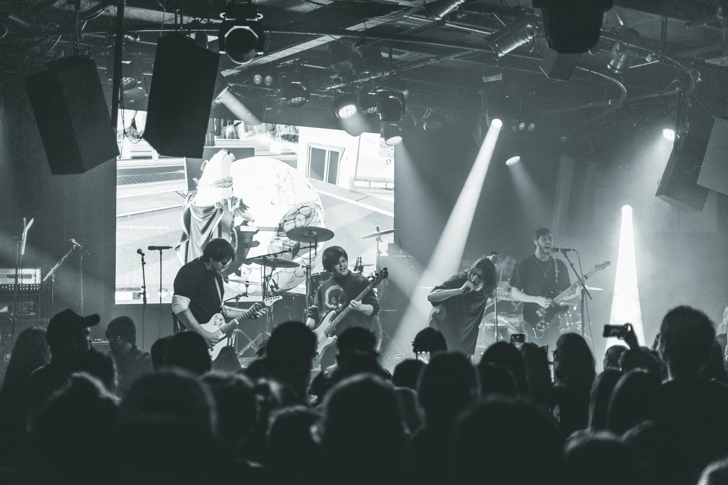 A live music performance with a band on stage, including guitarists, a vocalist, and a drummer, with lighting effects and an audience watching.