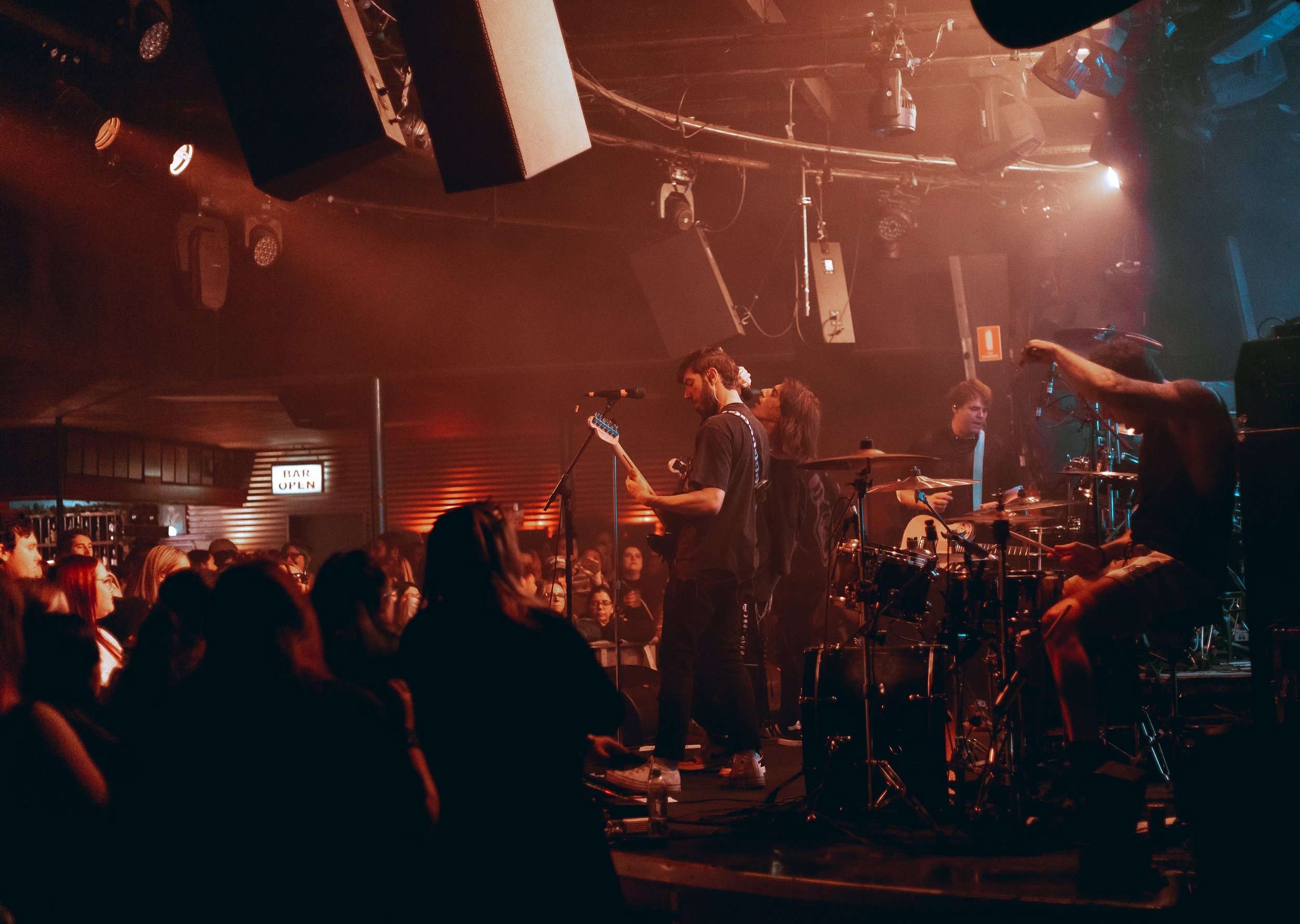 A live music band performing on stage in a dimly lit venue with an audience watching. The band includes a guitarist and a drummer, with various stage lights and equipment visible.