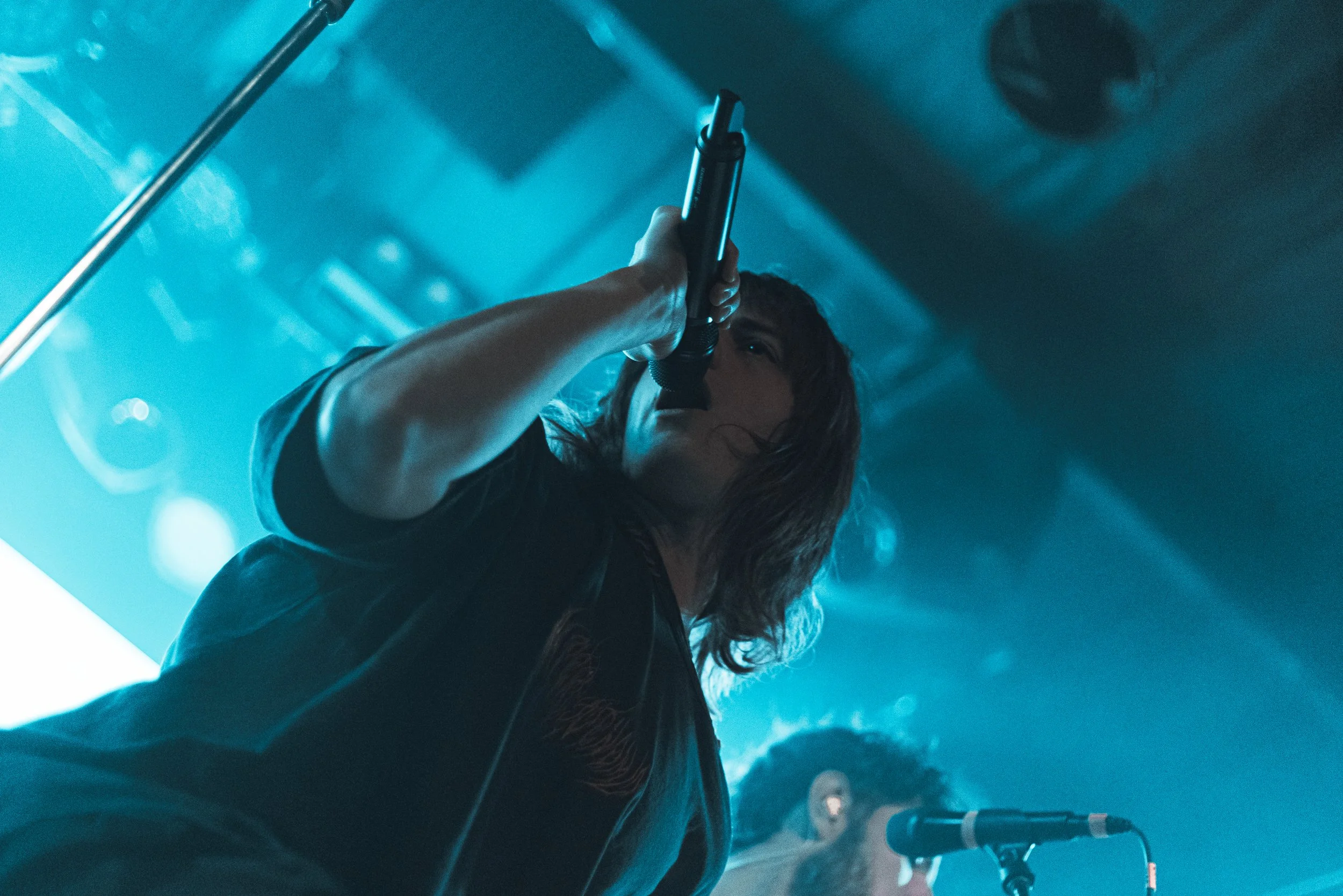 A person with long hair holding a microphone, performing on stage with blue lighting and fog effects.