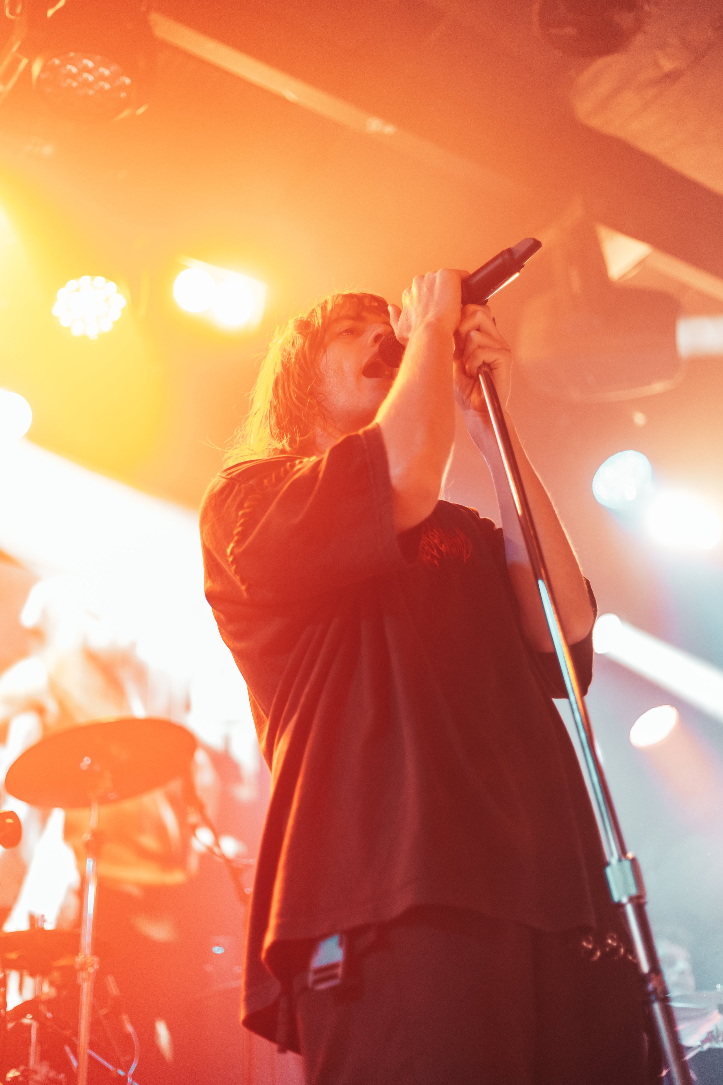 A male singer performing on stage, holding a microphone with both hands, with bright stage lights illuminating him from above and behind, creating a warm orange glow.