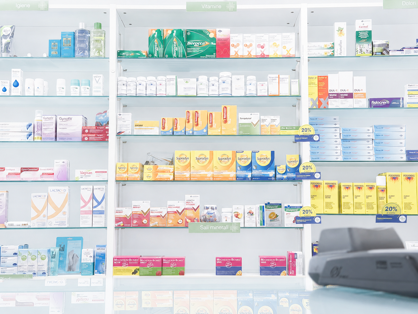 Scaffale di farmacia con vari farmaci e integratori, divisi per sezioni come igiene, vitamine, sali minerali e dolori.