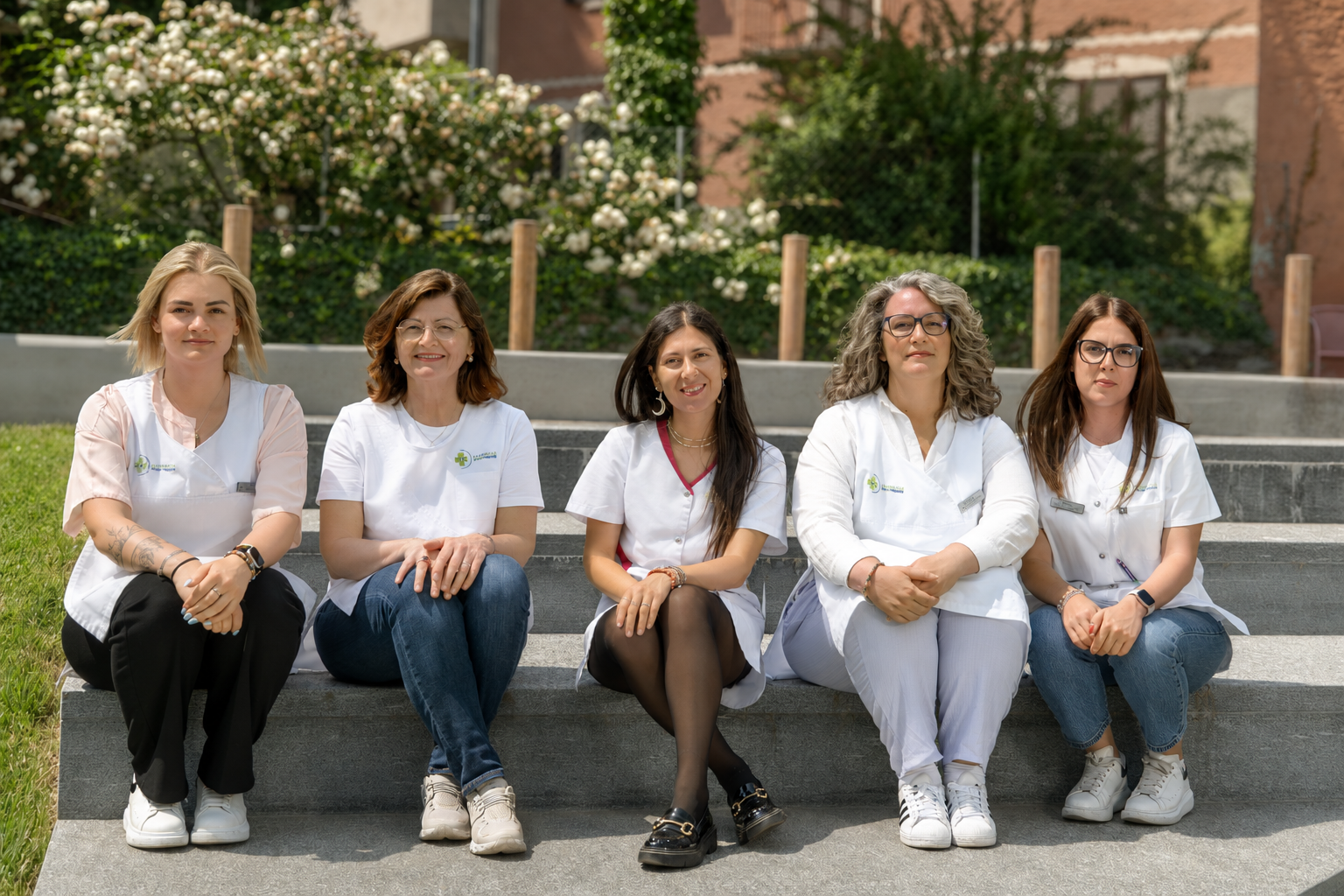 Cinque donne sedute su gradini all'aperto in una giornata di sole, con piante e fiori sullo sfondo, indossando abbigliamento bianco e sorridendo.