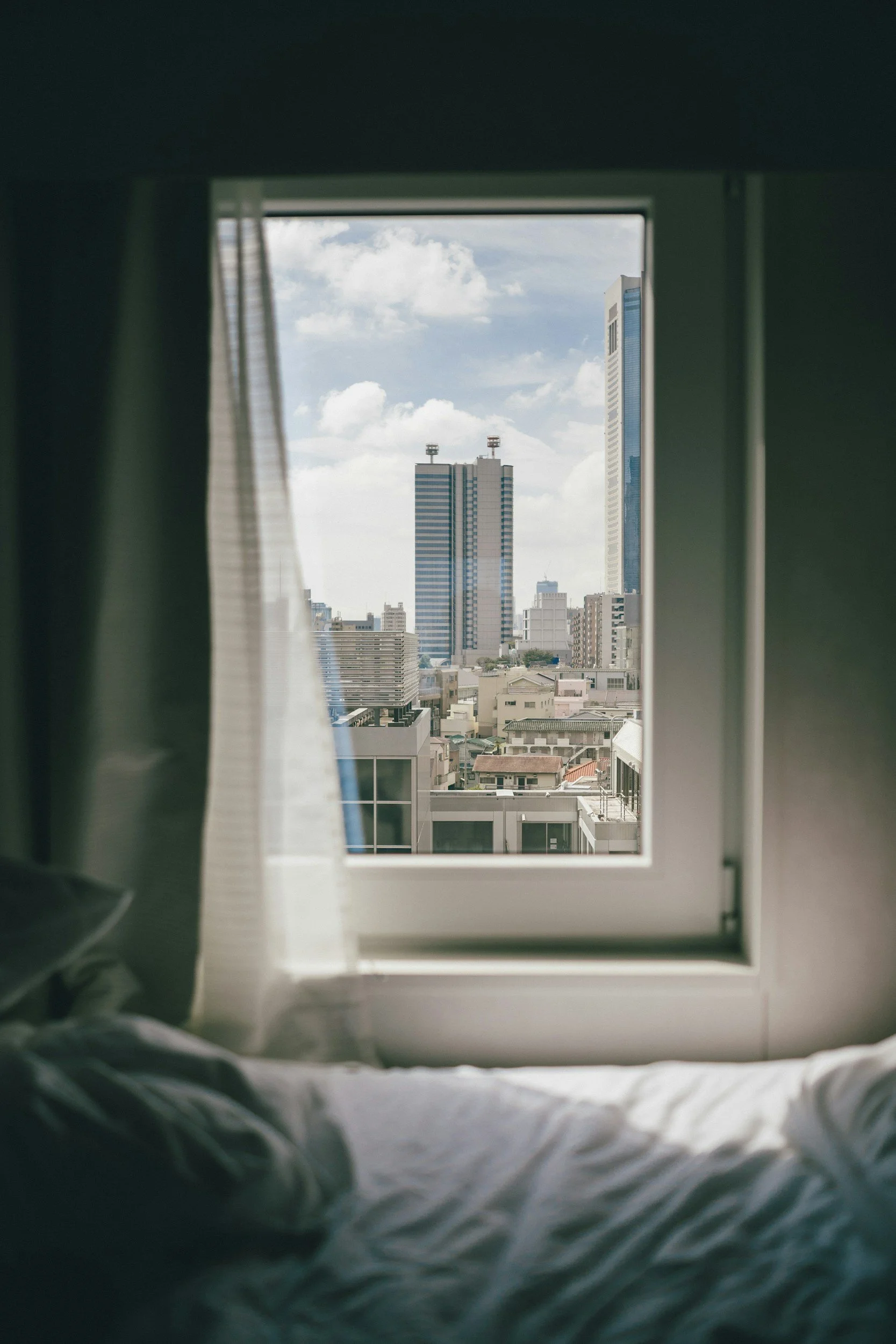 View through a window showing city skyscrapers and a cloudy sky, framed by curtains and a bed with sheets in the foreground.