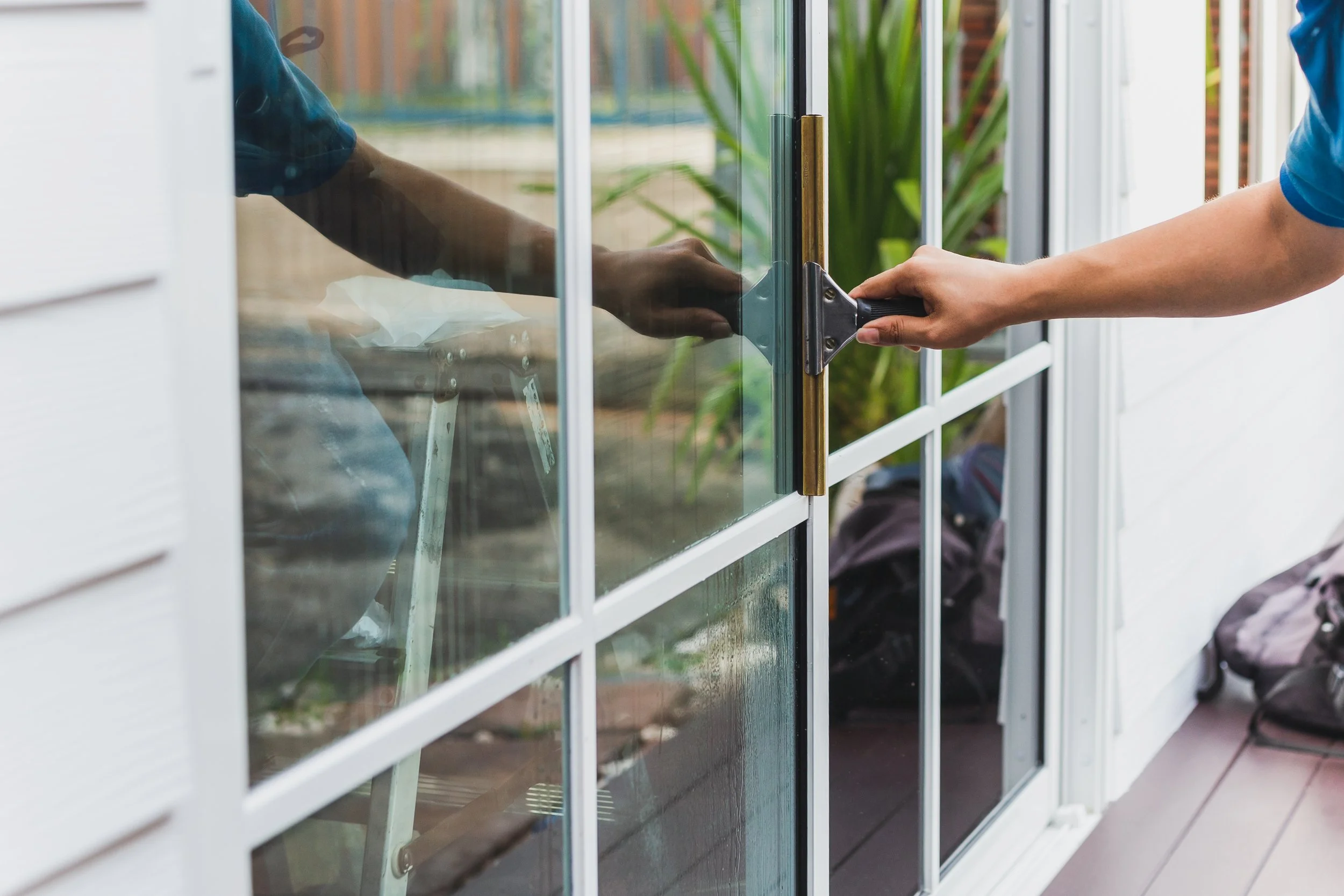 A person in a blue shirt opening or closing a glass door with a black handle and a vertical brass strip. The glass door reflects greenery from outside, and a person with a backpack can be seen sitting inside.