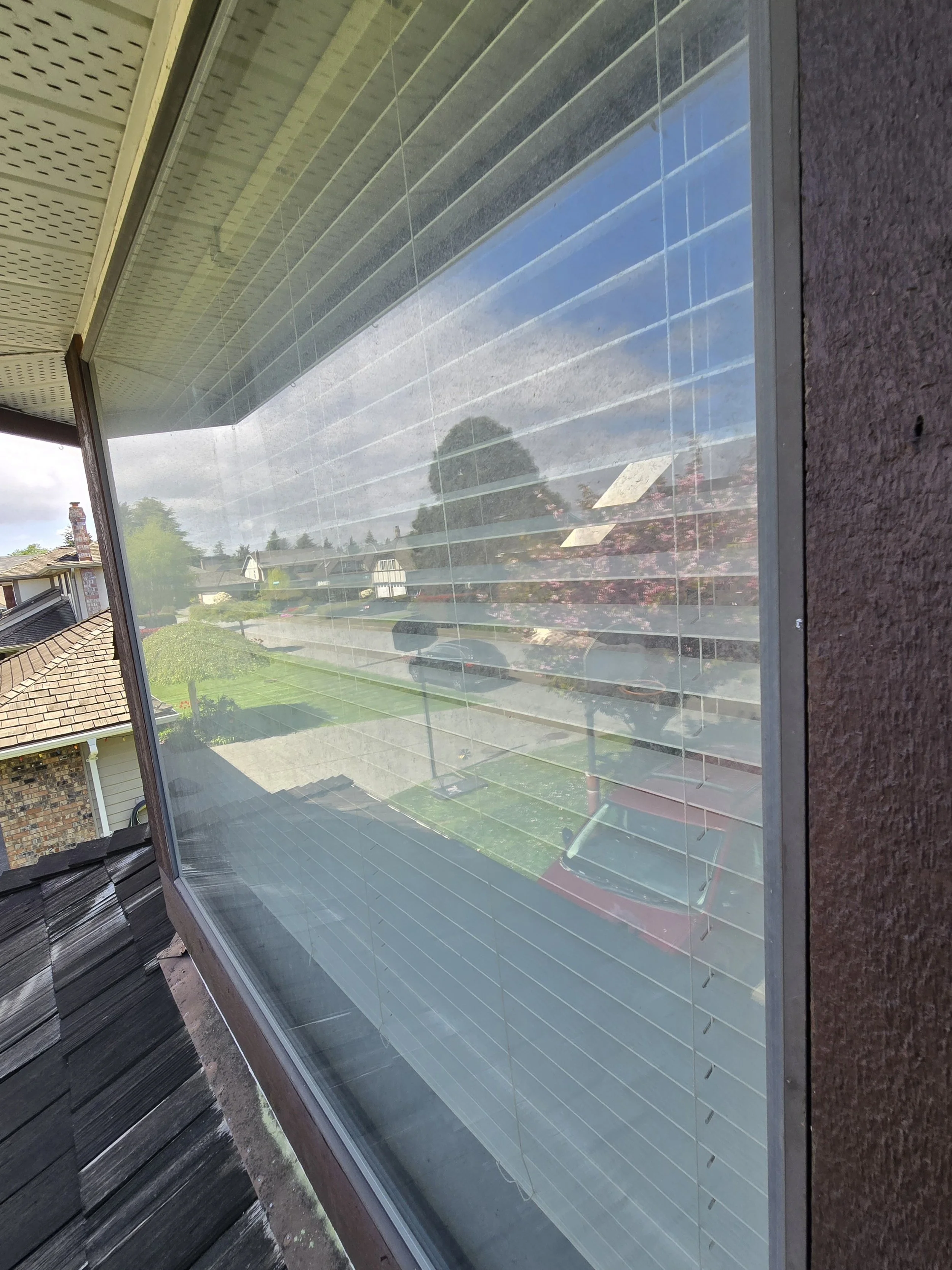 A window with horizontal blinds reflecting a suburban neighborhood scene, including houses, trees, and a lawn, with an outdoor overhang and roof shingles visible.