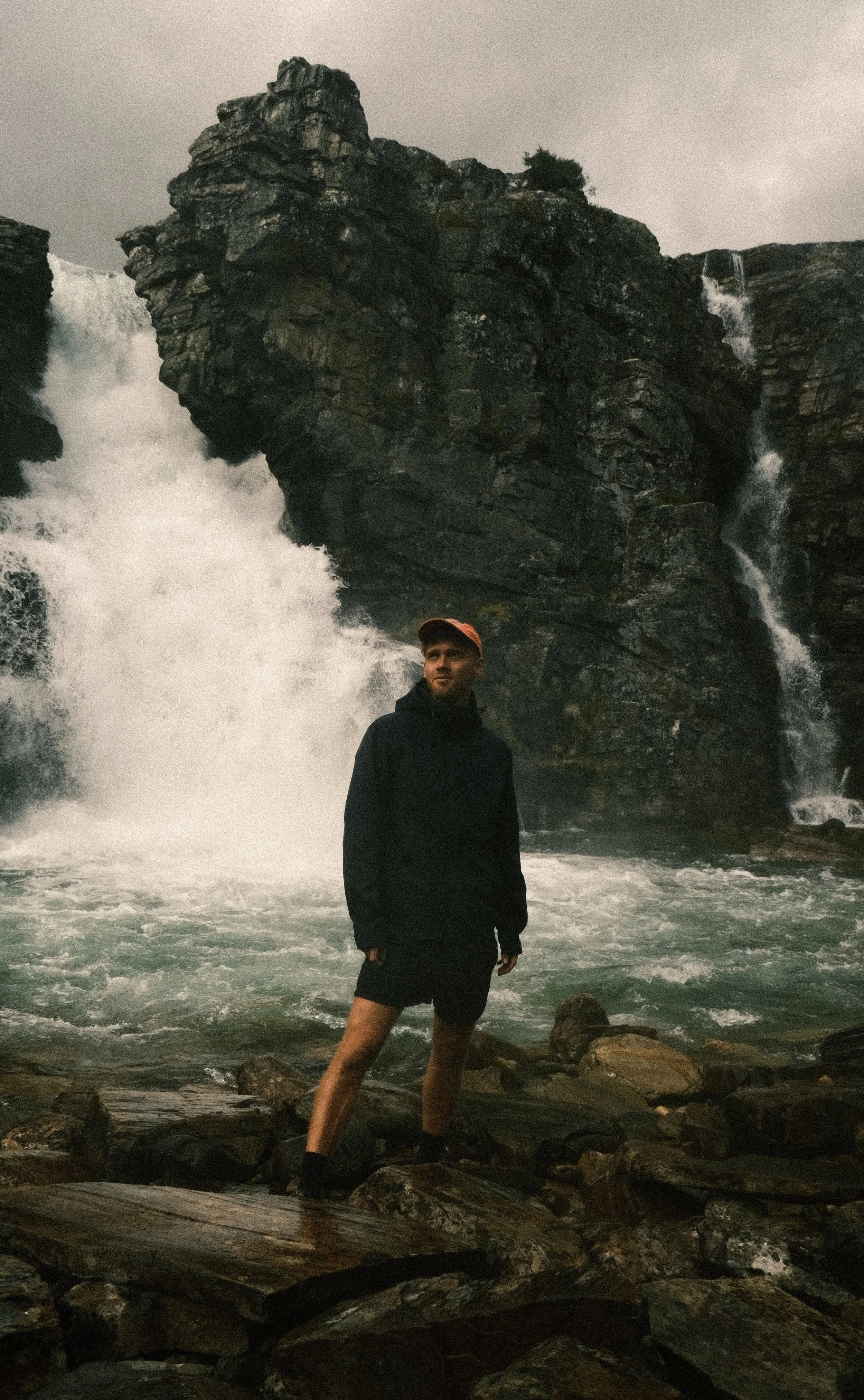 A person standing on rocks at the edge of a waterfall with large rock formations and rushing water in the background.