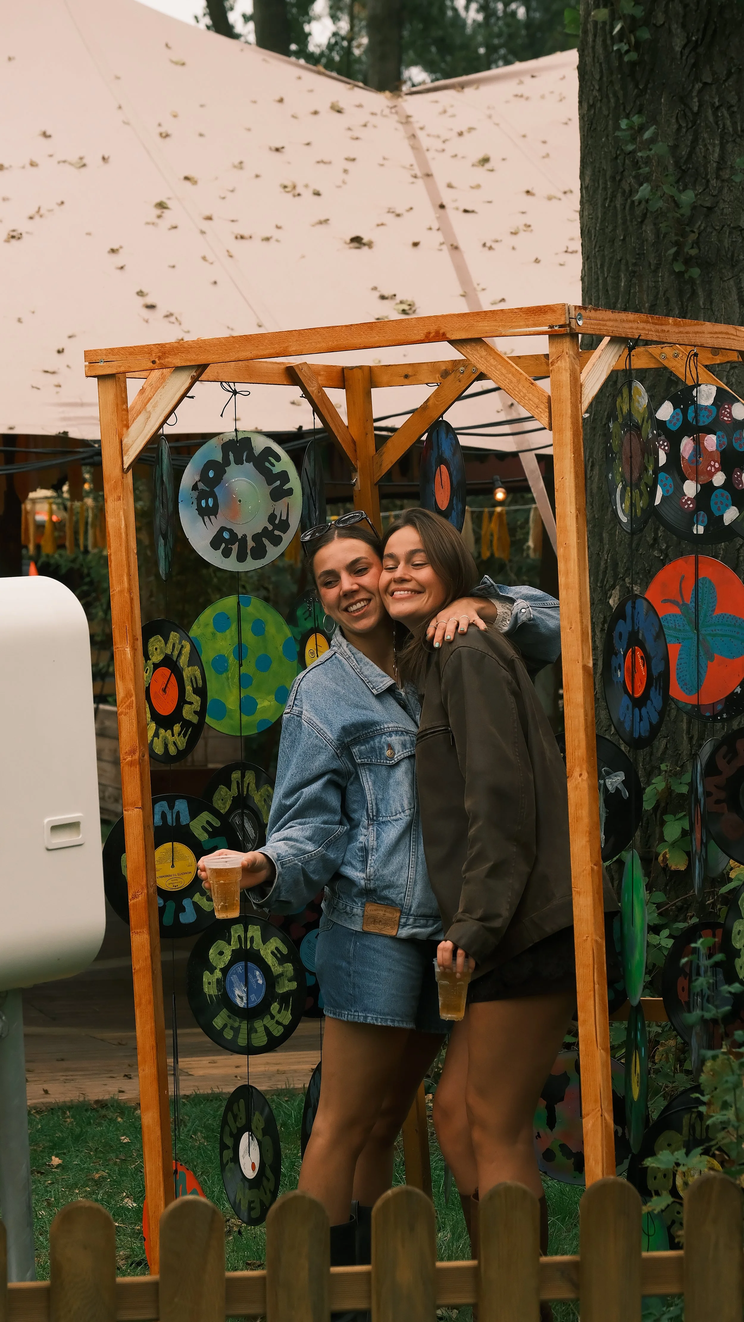Twee vrouwen die lachen en elkaar omhelzen binnen een houten fotomuur met kleurrijke kunstwerken, met een tent en bomen op de achtergrond.