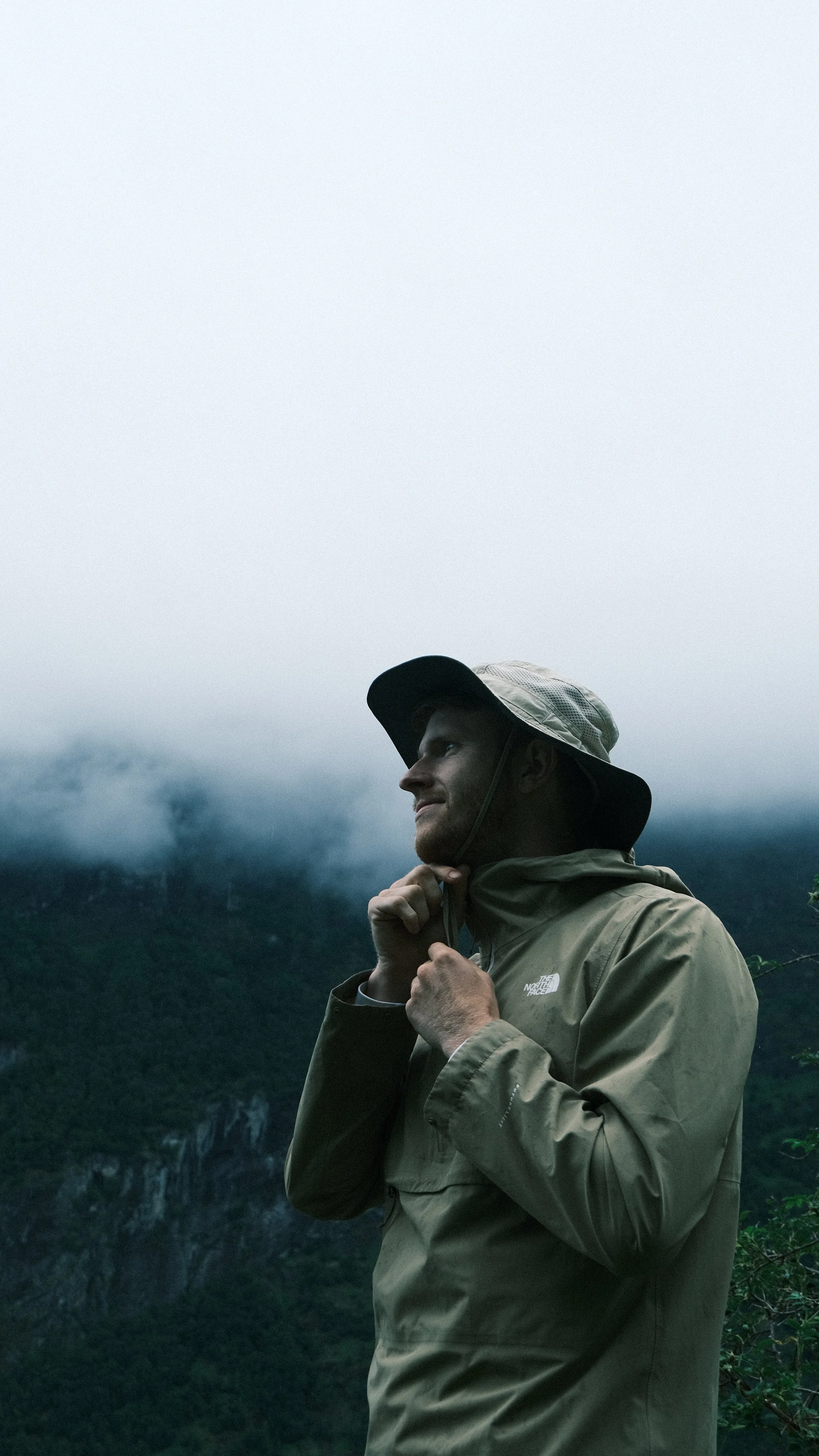 Man in een beige jas en hoed, staand in een bergachtig landschap met mist, kijkt tevreden en denkt na.