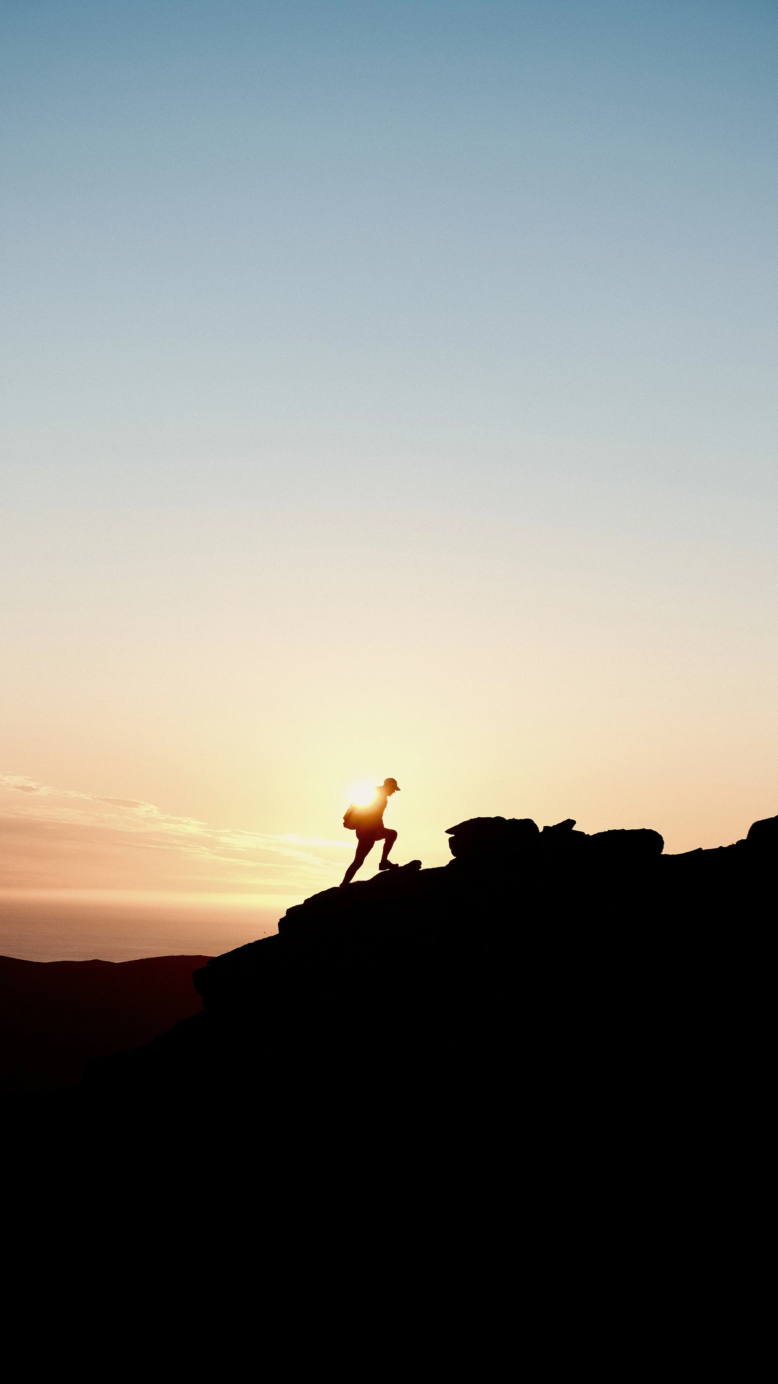 A person hiking on a mountain at sunset with the sun behind them, silhouetted against a colorful sky.