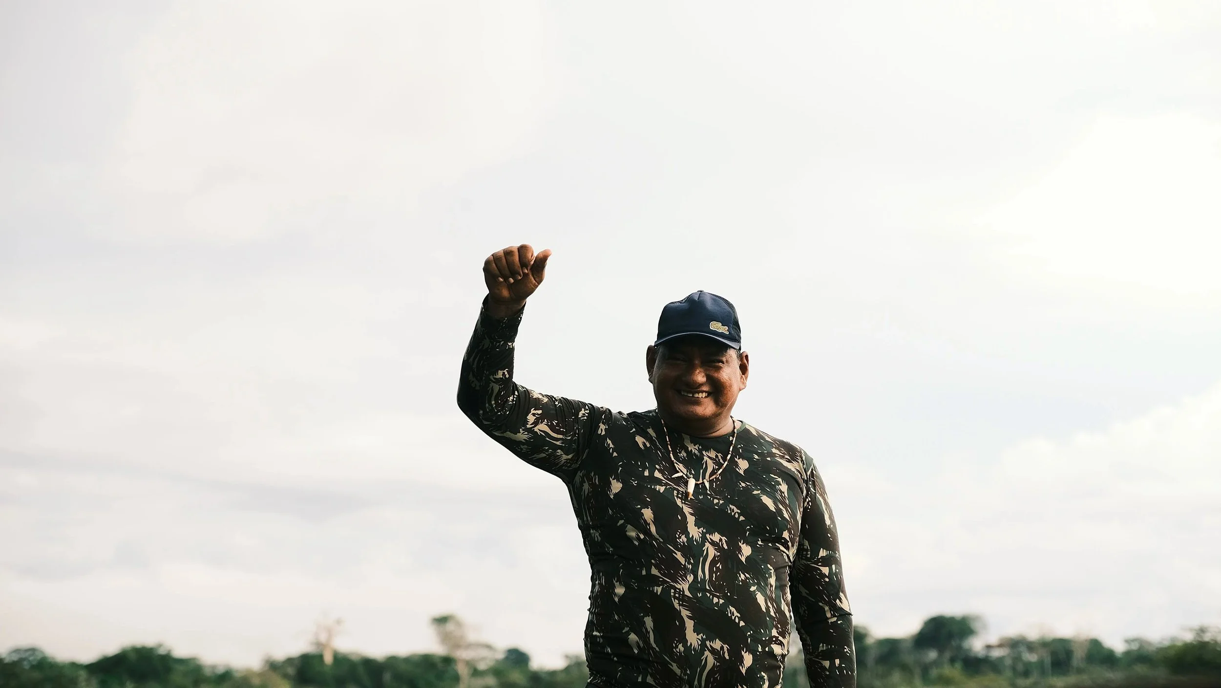 Vrolijke man in camouflageprint shirt en pet, zwaaiend met zijn hand, buiten onder een bewolkte hemel.