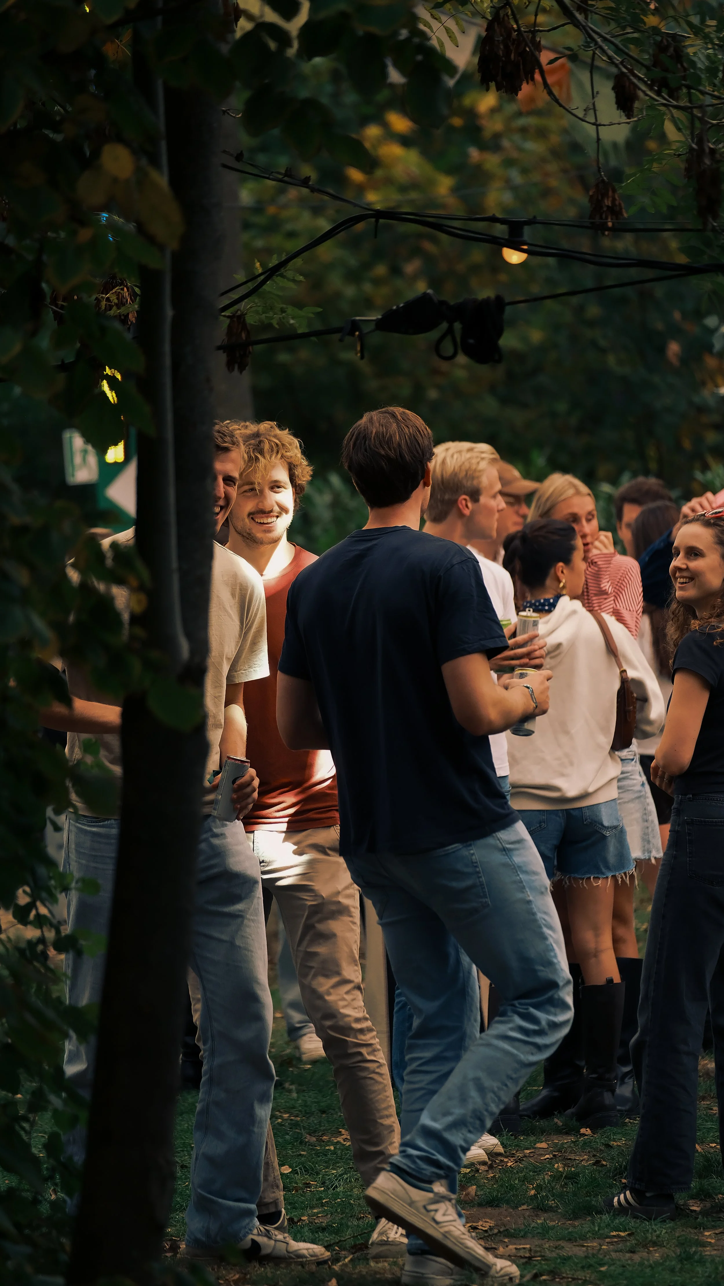 Groep jongeren die samen plezier hebben in een park 's avonds, met bomen en bladeren op de achtergrond en warme verlichting.