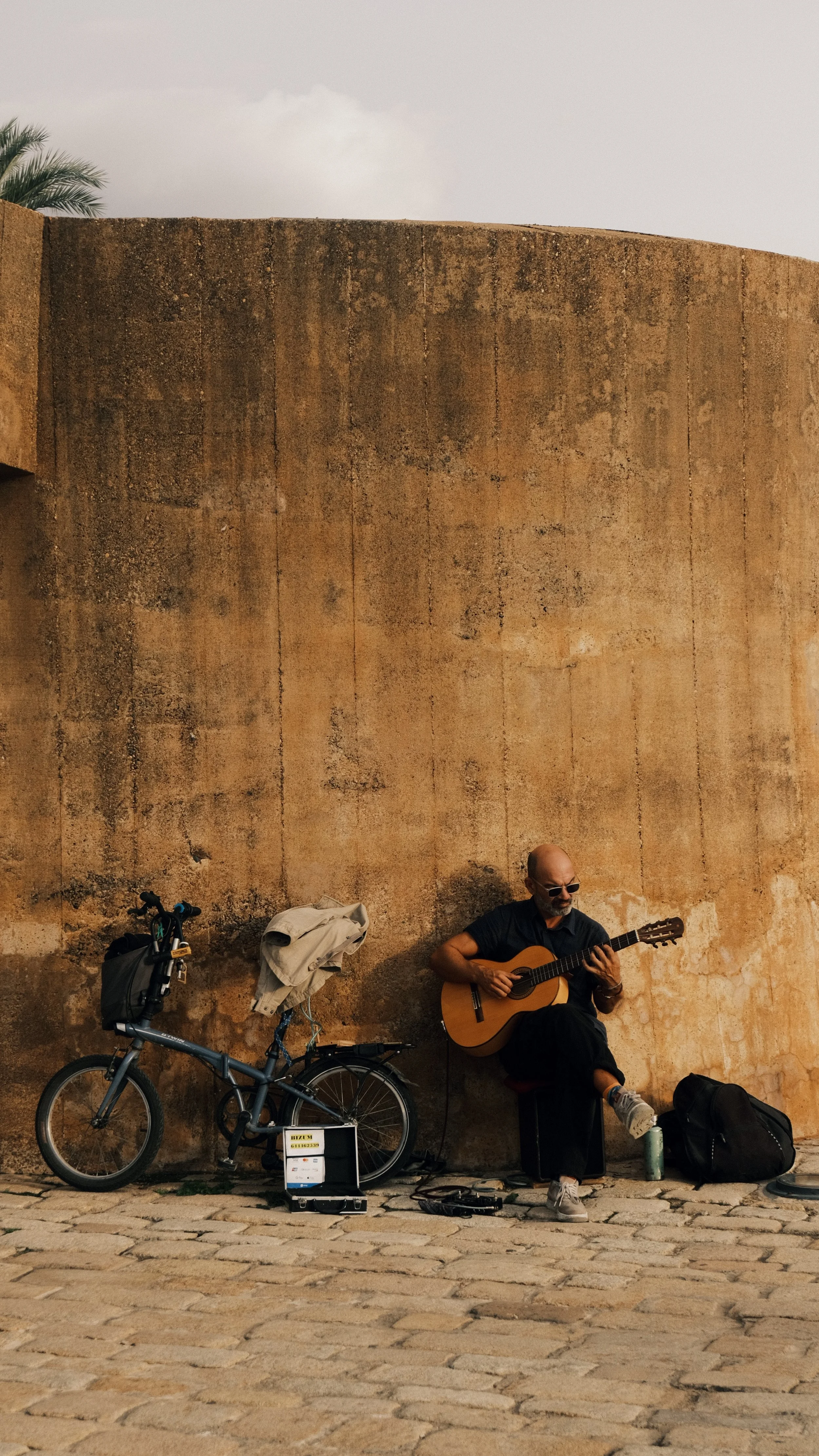 Man speelt gitaar terwijl hij op een bankje eindigt tegen een grote stenen muur. Er is een fiets, een doos met spullen en een rugzak naast hem.