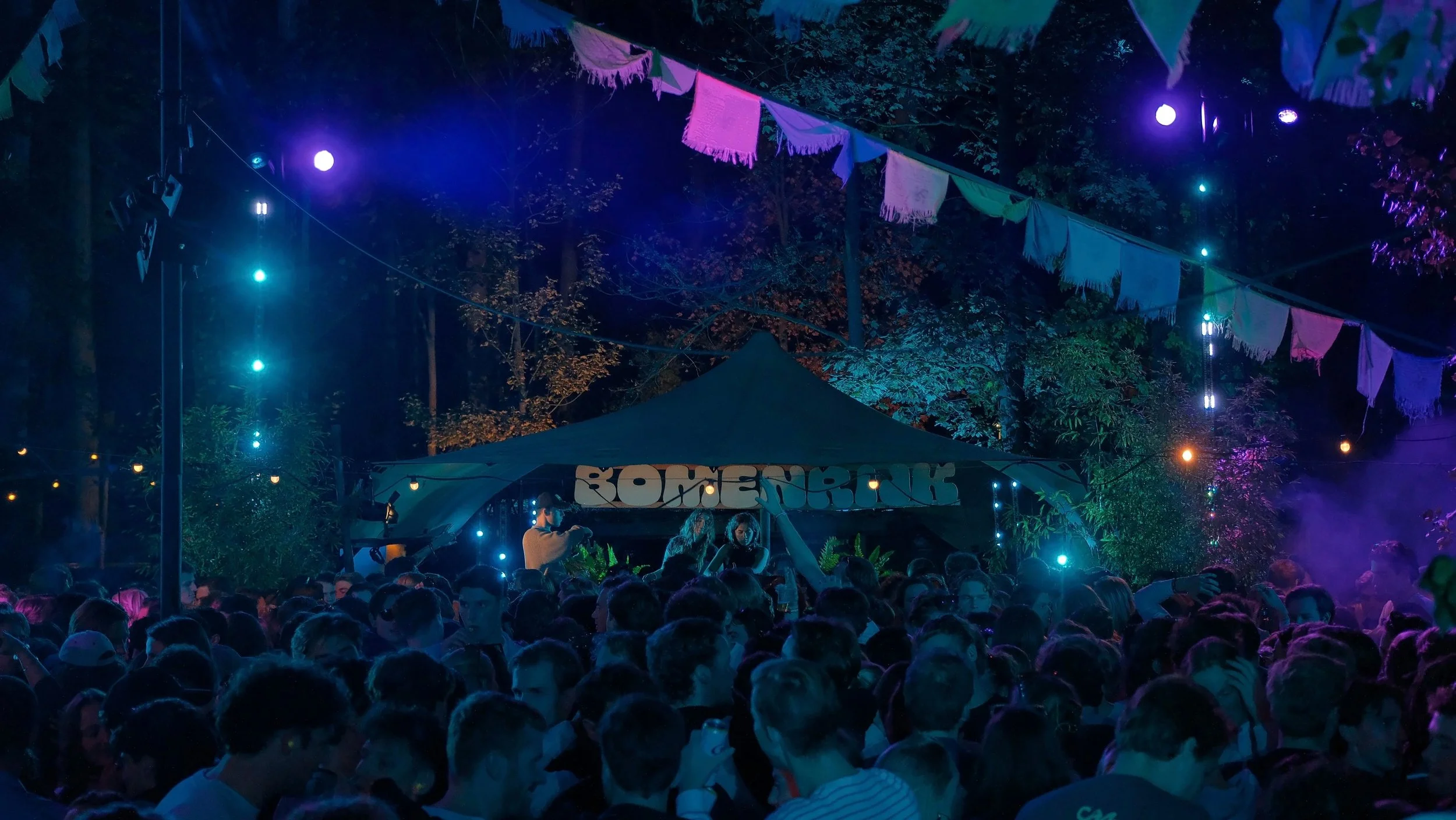 Nighttime outdoor festival scene with colorful string lights and bunting decorations. A dense crowd of people is gathered, and a stage with a tent displaying the word 'ROMER' is visible in the background. The scene is illuminated by various colored lights, creating a vibrant and lively atmosphere.