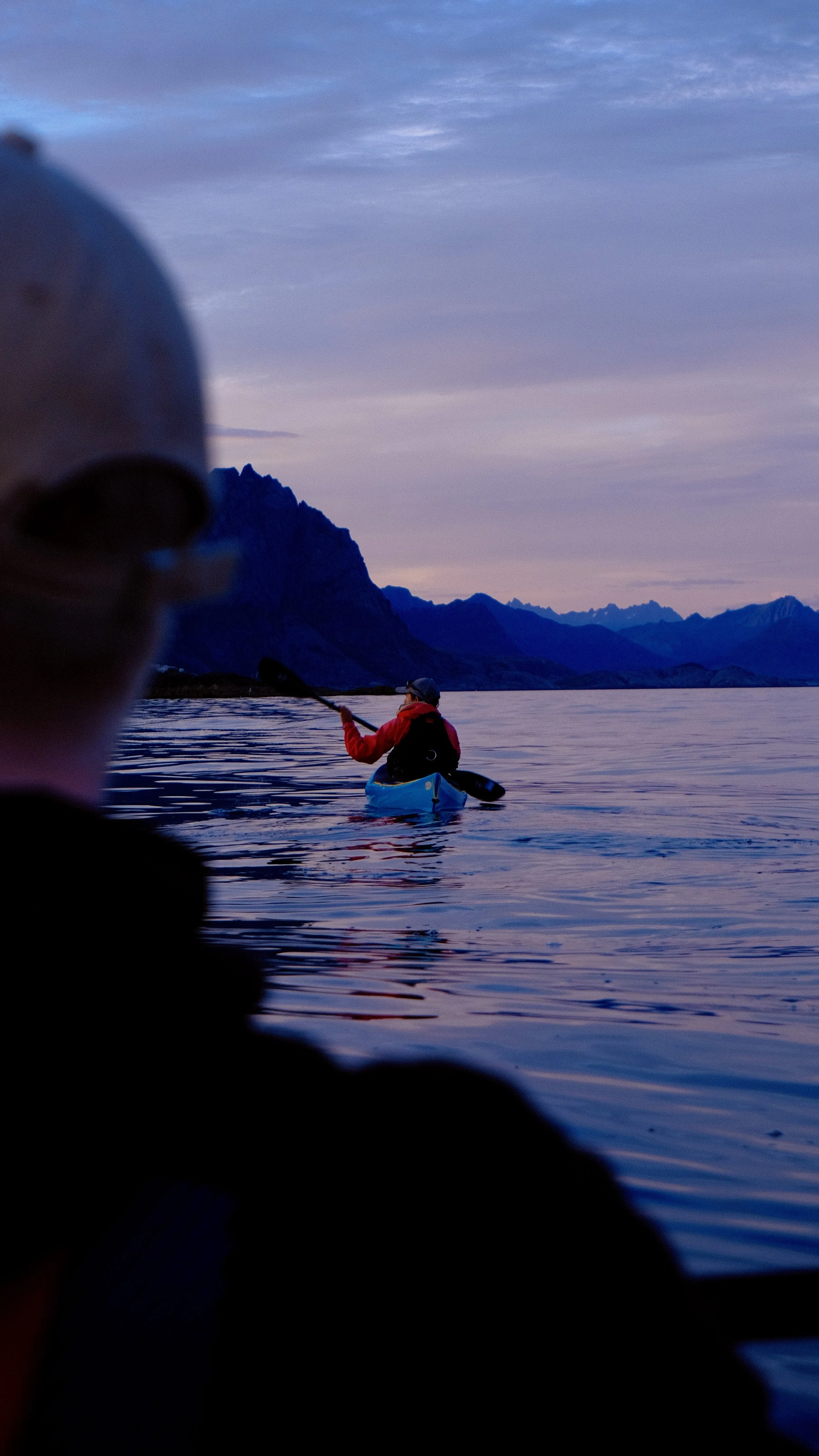 Een persoon in een rode jas en pet peddelt in een blauwe kajak op een kalm meer met bergen op de achtergrond tijdens zonsondergang.