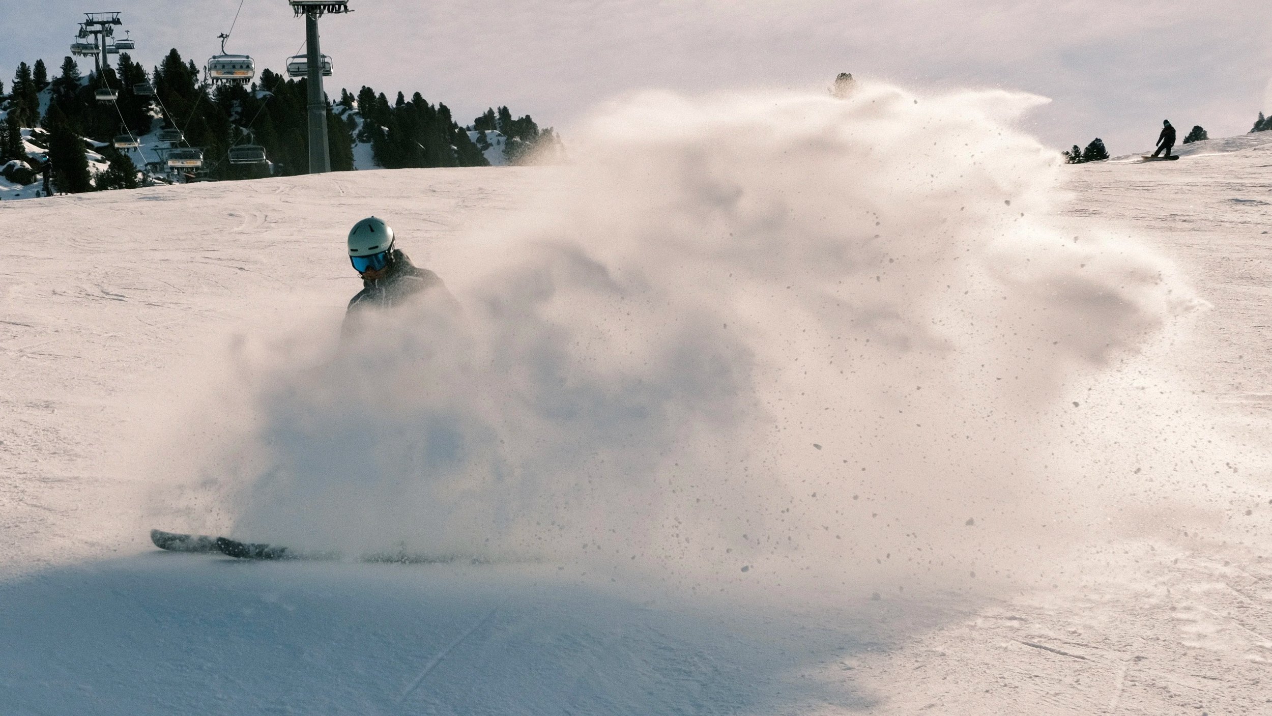 Sneeuwsporter die door de sneeuw rijdt op een skipiste, met een skibril en helm, terwijl er skiliften en andere skiërs op de achtergrond zijn.