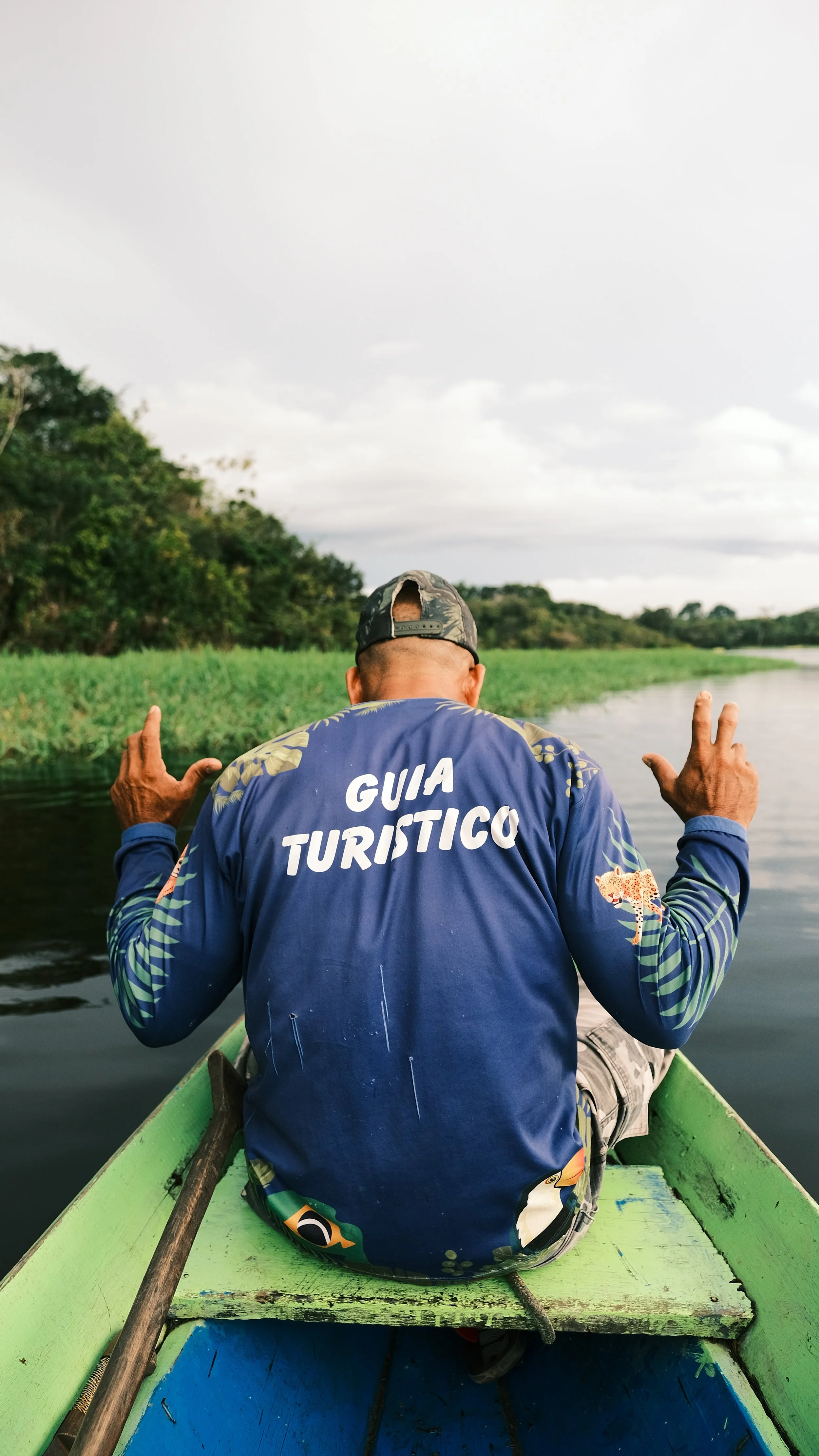A man wearing a blue shirt with the words 'Guía Turístico' on the back is sitting in a green boat on a river, surrounded by lush green trees and an overcast sky.