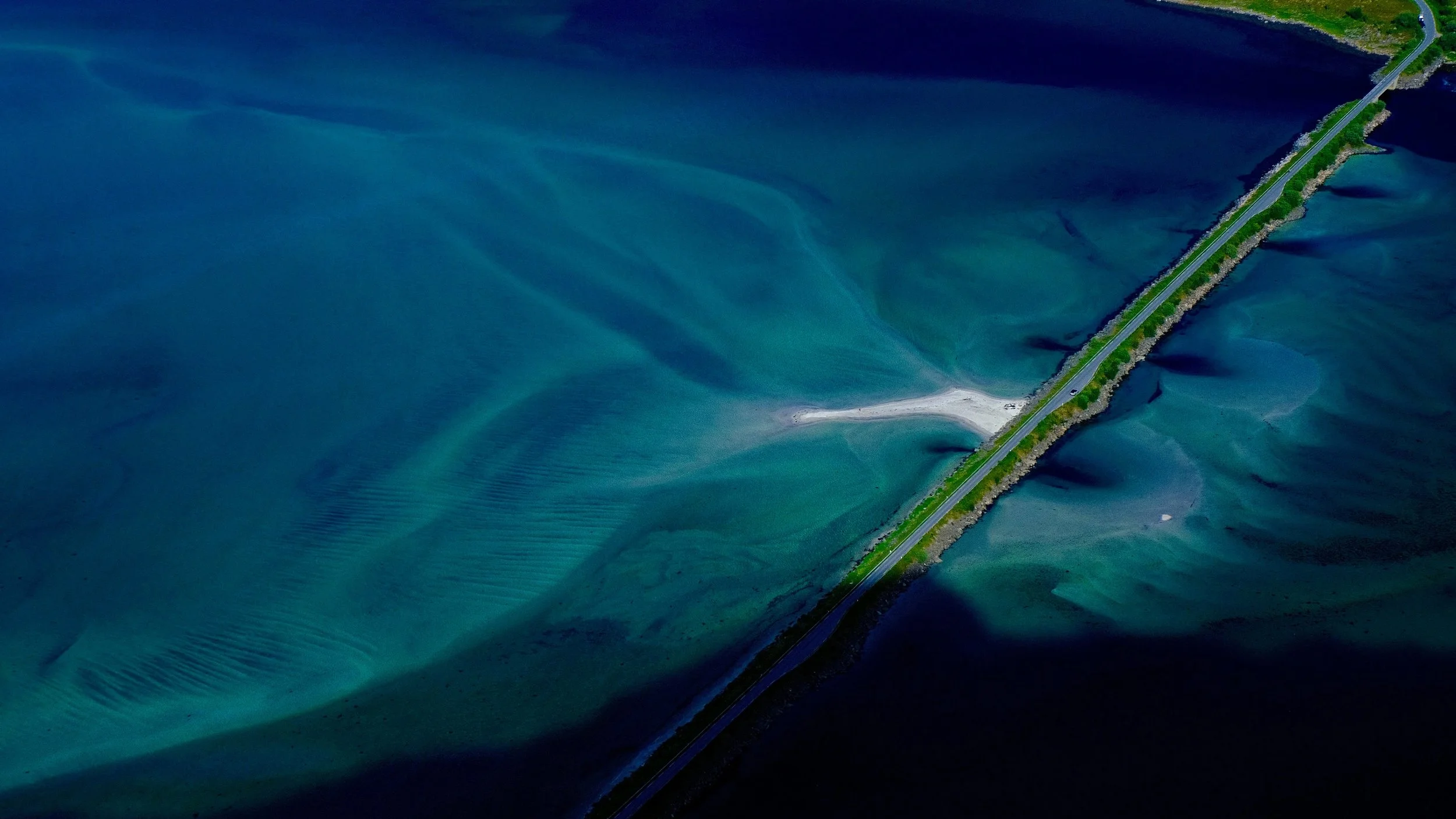 Aerial view of a dam or bridge over a body of water with varying shades of blue and green, with a road running along the top.
