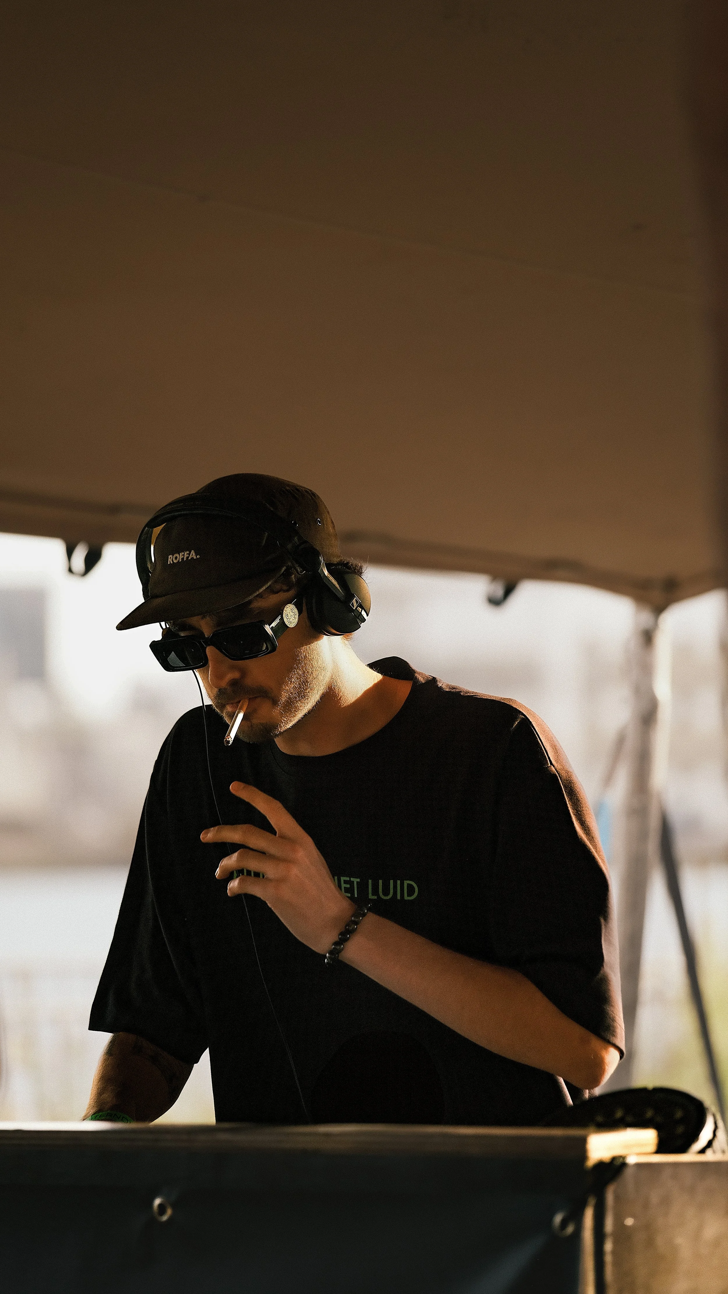 Man met een pet, zonnebril en hoofdtelefoon onder een tent, rookt een sigaret en staat achter een dj-setup.