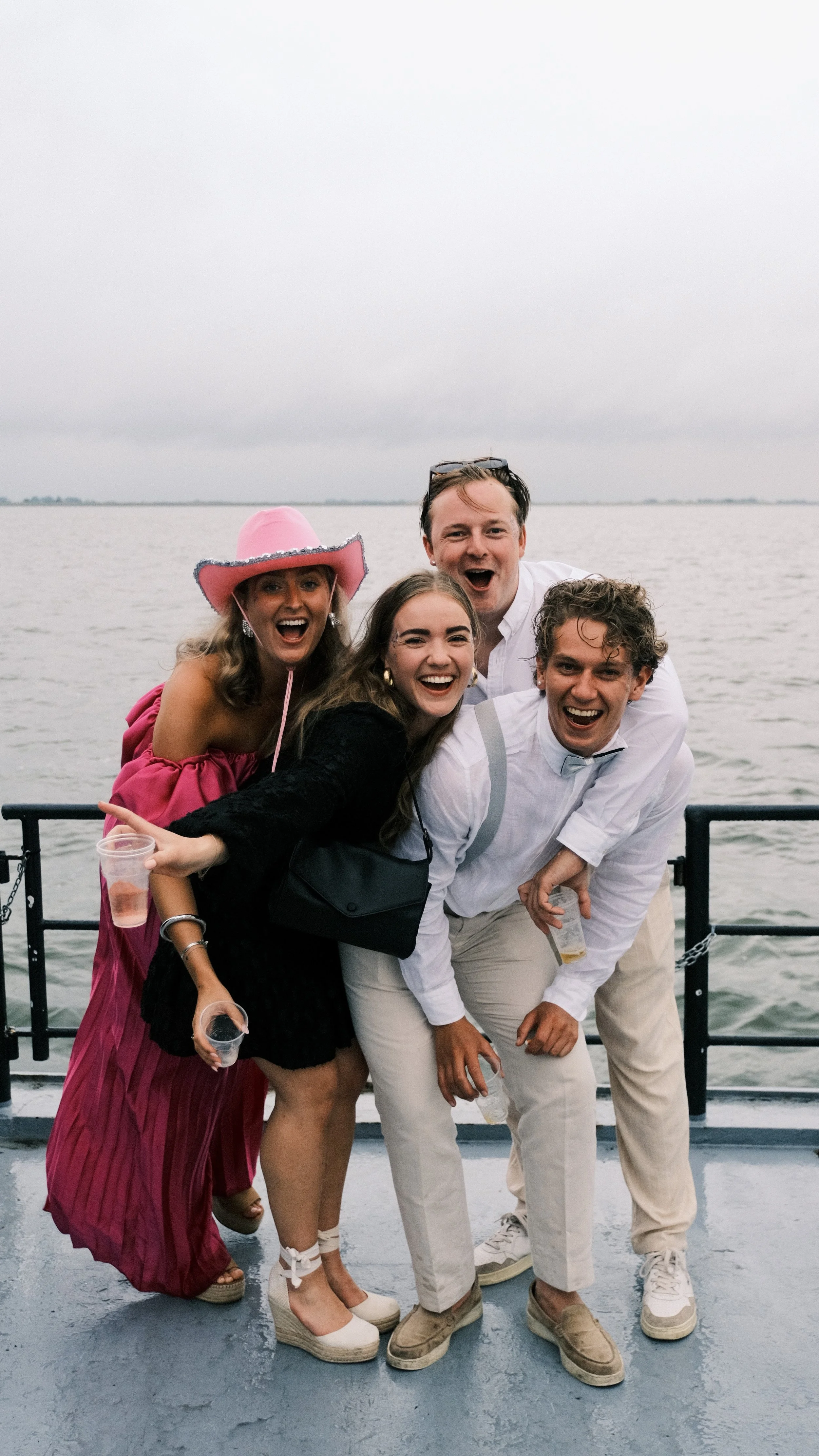Vijf mensen die vrolijk lachen op een boot met water op de achtergrond, onder een grijze hemel.