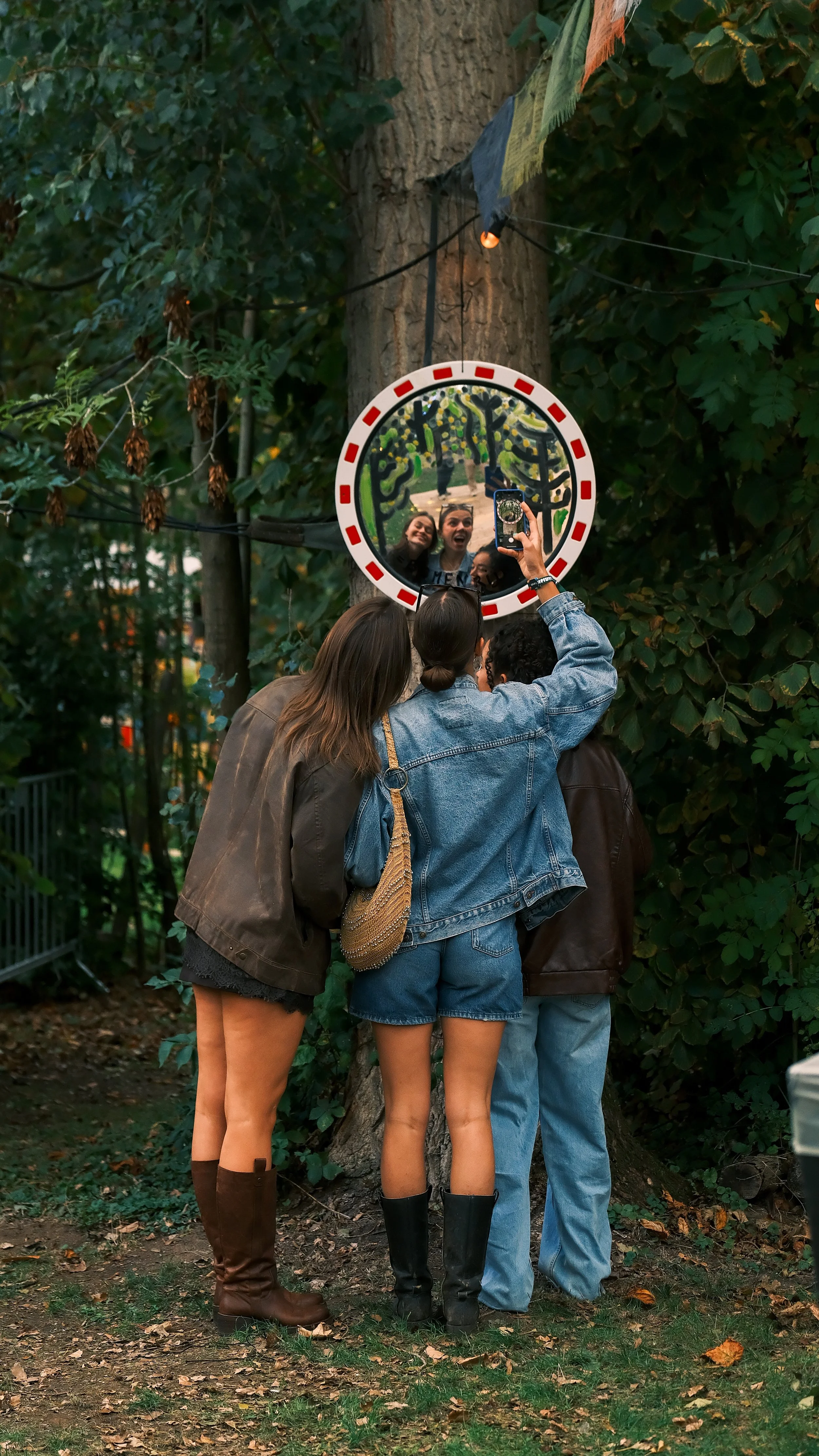 Vrouwen kijken in een ronde spiegel die aan een boom is bevestigd, omarmen elkaar en maken een foto met een smartphone in een bosrijke omgeving.