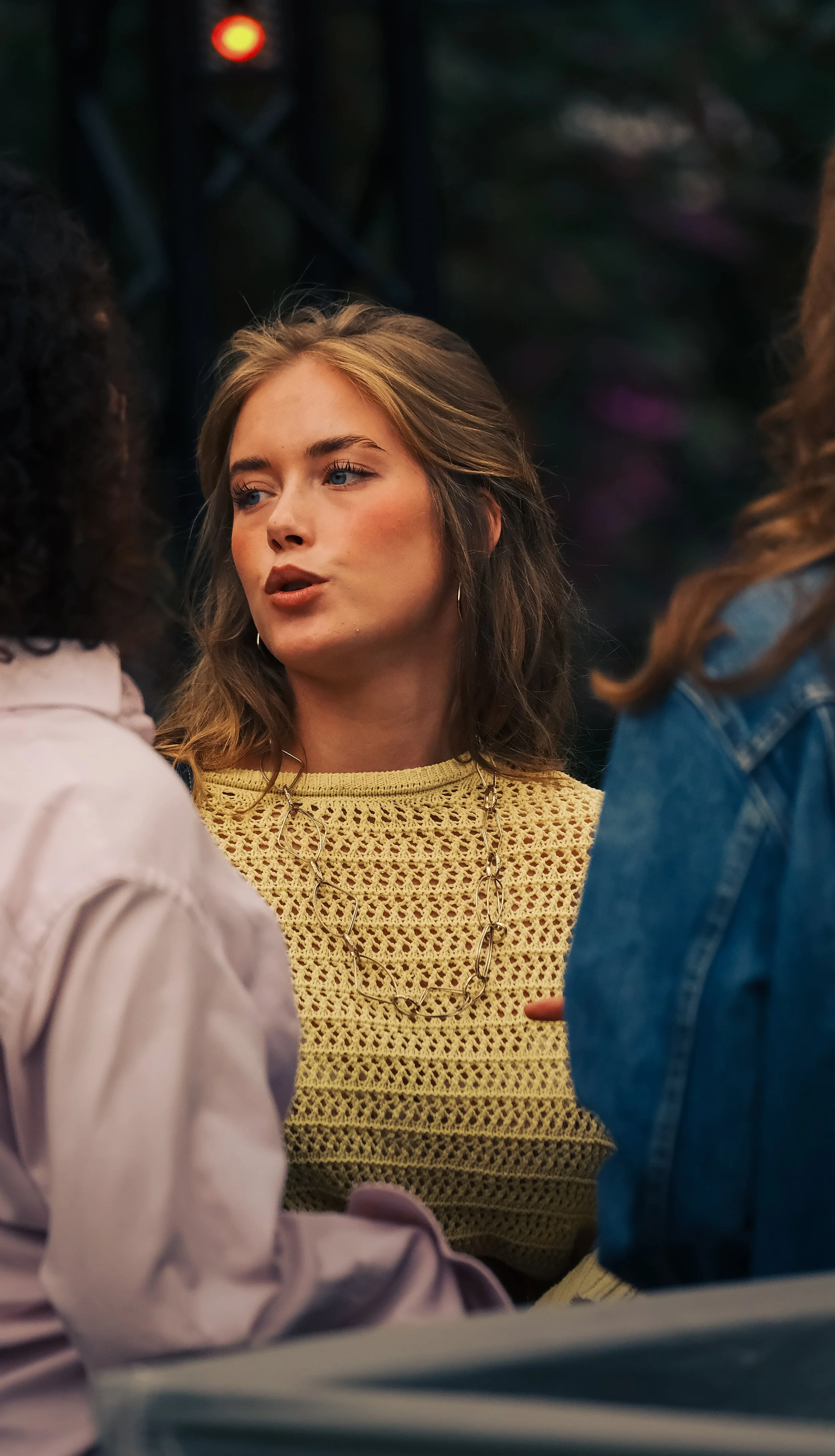 Een jonge vrouw met blond haar en blauwe ogen, gekleed in een geel gebreid top en een ketting, spreekt met anderen.
