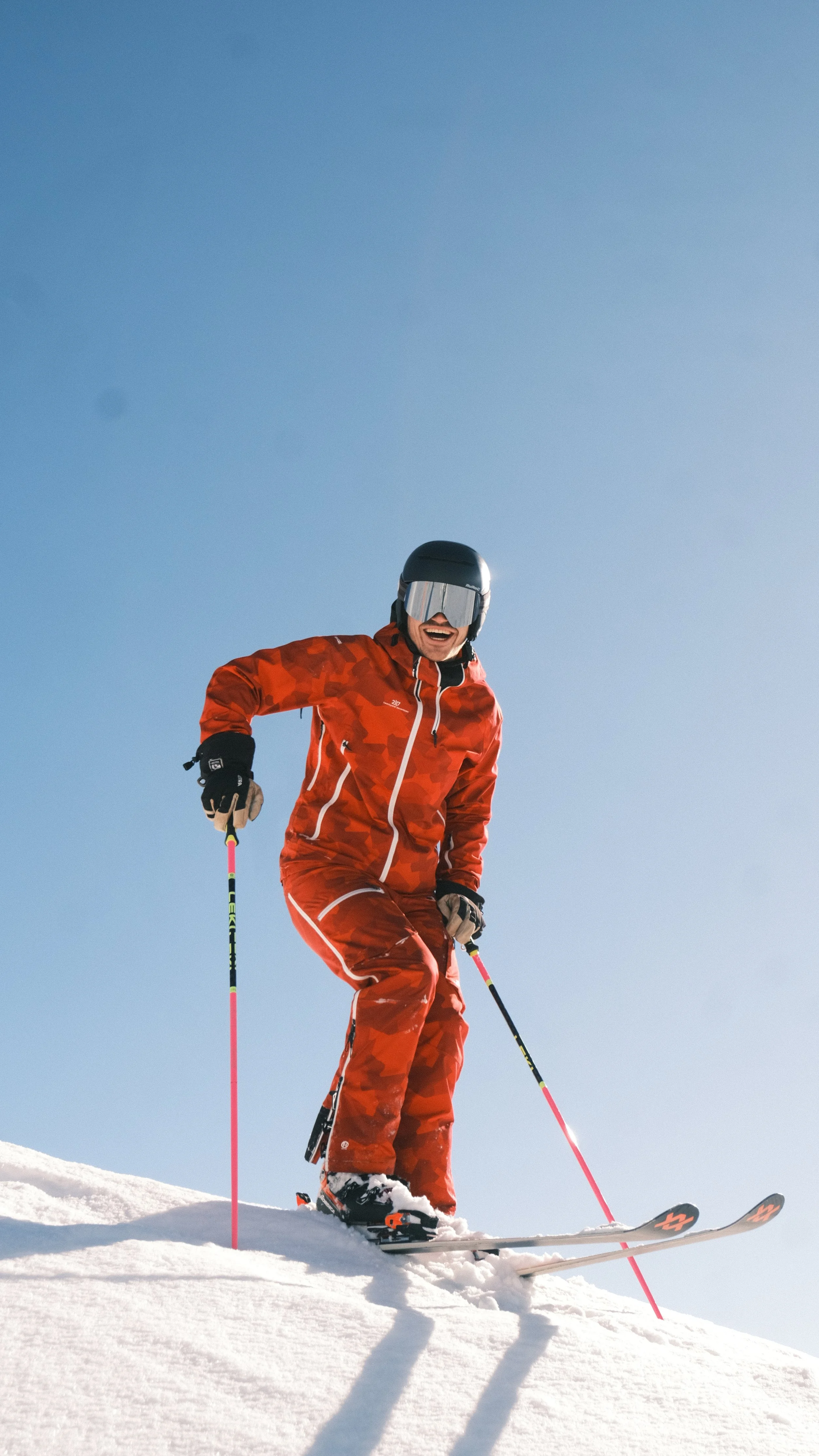 Een lachende skiër in een rood oranje skipak met een helm en skibril, skiet op een besneeuwde berg onder een heldere blauwe hemel.