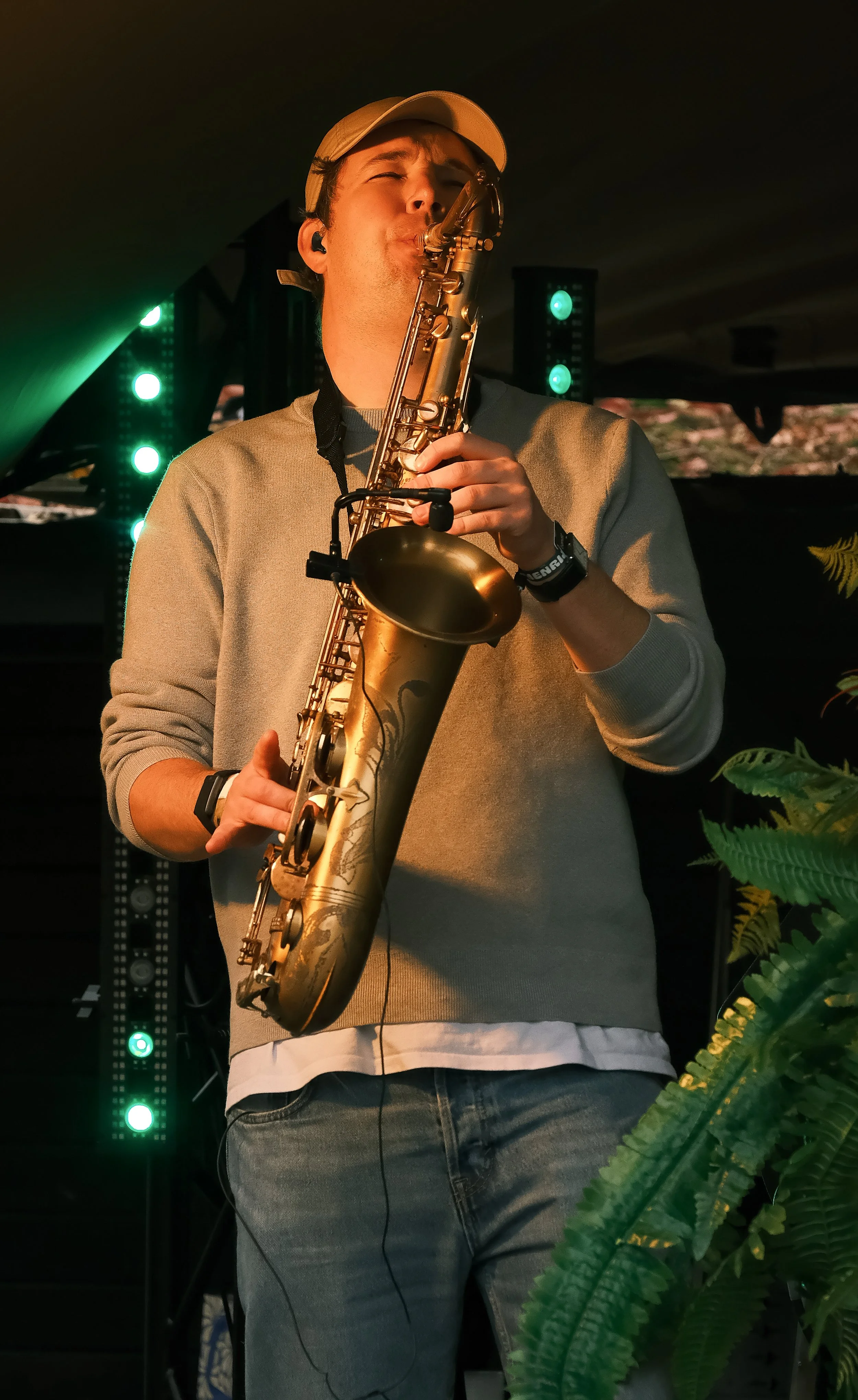 A man wearing a beige cap and light gray sweater playing a gold saxophone on stage, with green stage lights and foliage in the background.
