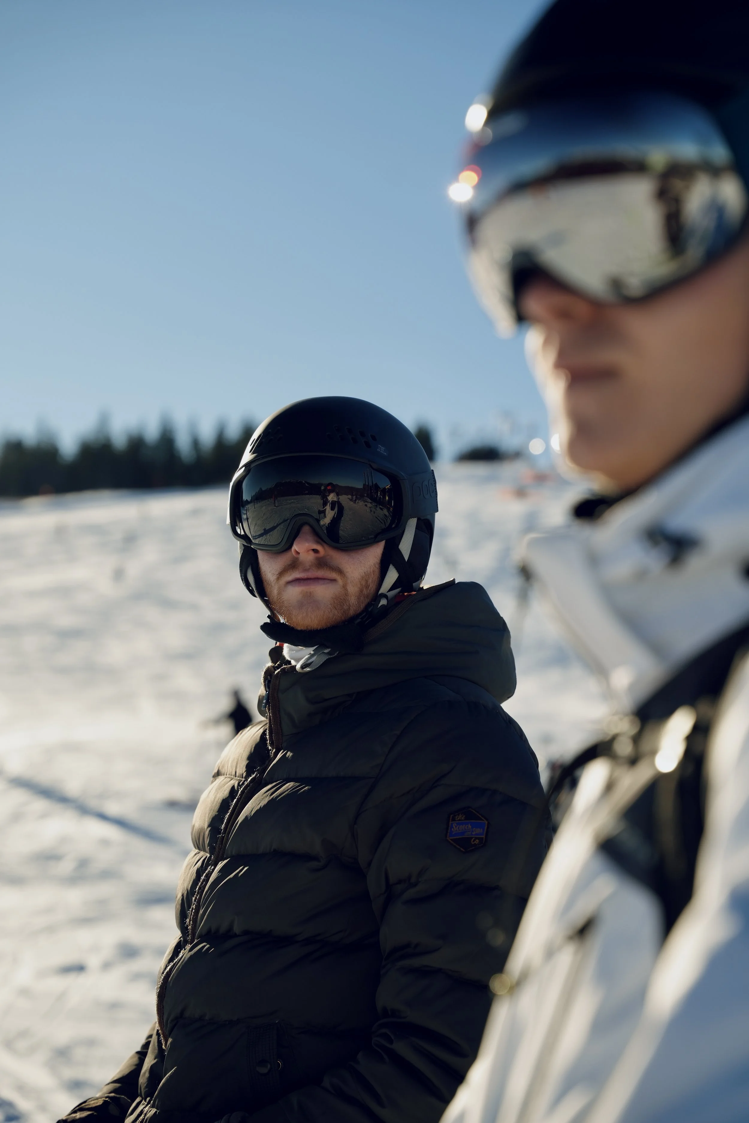 Two people in winter gear and helmets, wearing sunglasses, stand outdoors in a snowy landscape with a clear blue sky, with trees in the background. The person in the foreground is wearing a black jacket and the person in the background is wearing a white jacket.