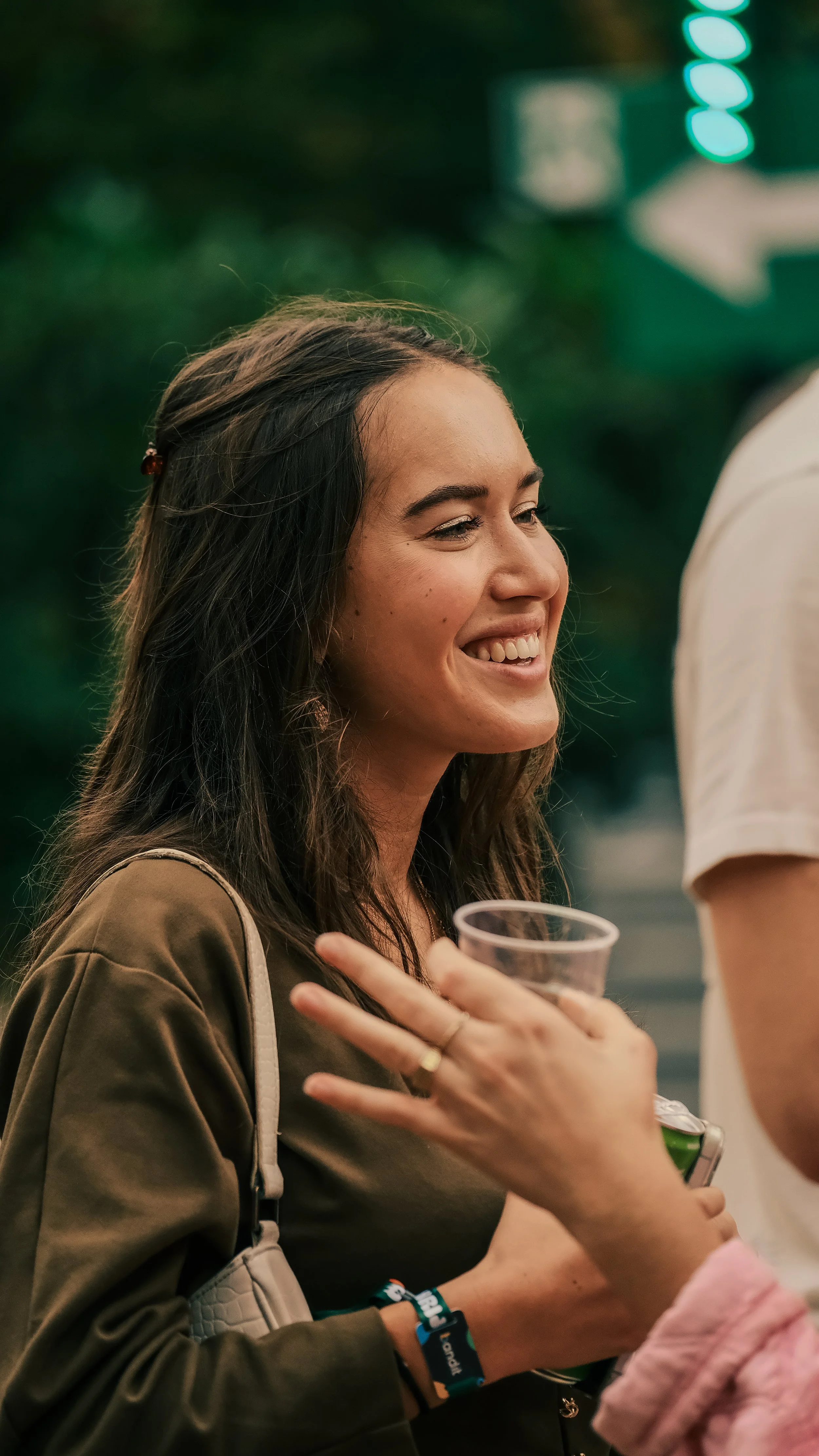 Vrouw met lang donker haar glimlacht terwijl ze in een plastic beker drinkt tijdens een sociale gebeurtenis, omgeven door andere mensen in een buitenomgeving.