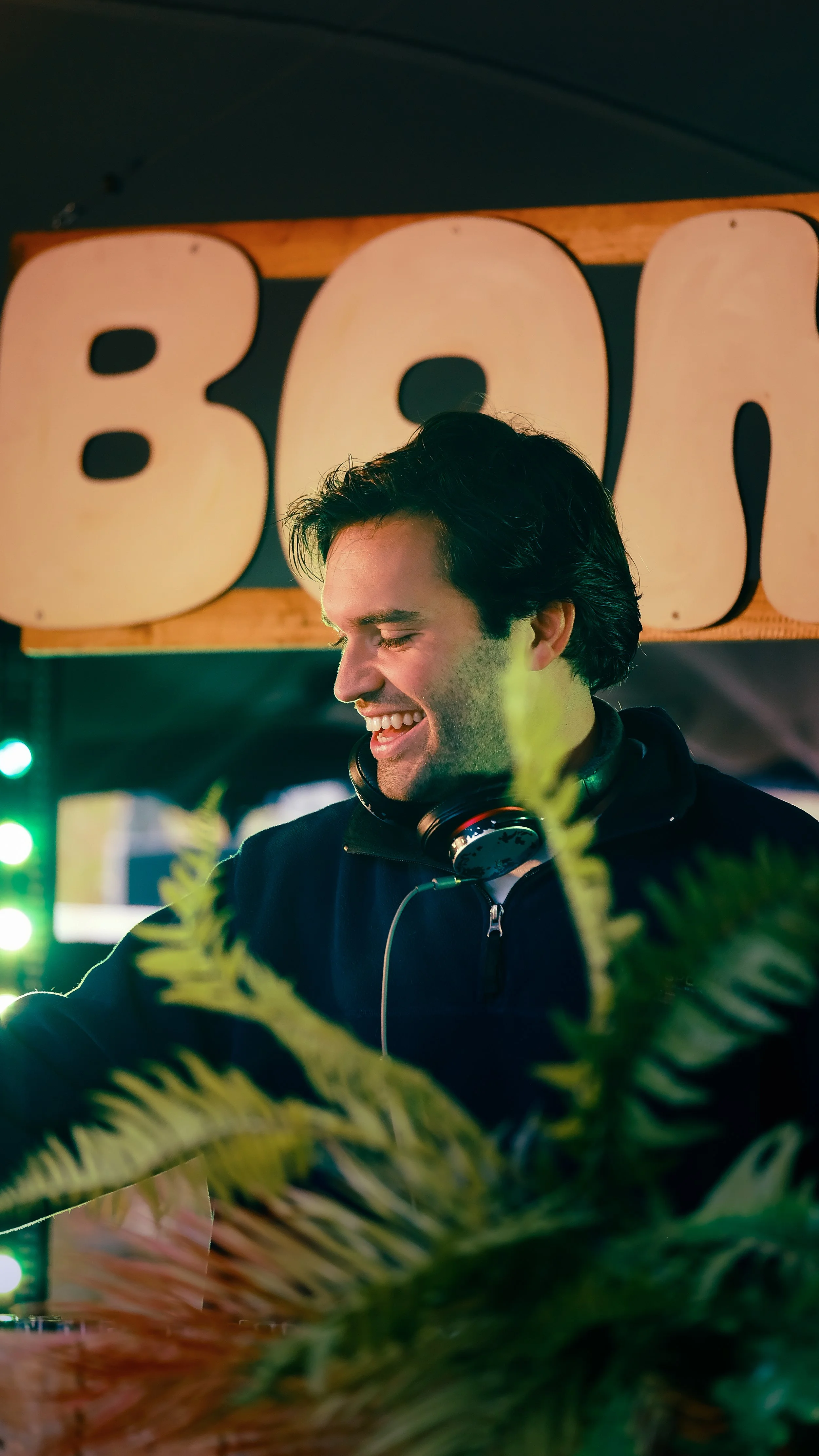 Een lachende man met zwarte haar, draag een zwart jack en hoofdlovens, staat voor een grote borden met de letters 'BOH', omringd door groene planten en gekleurd licht op de achtergrond.