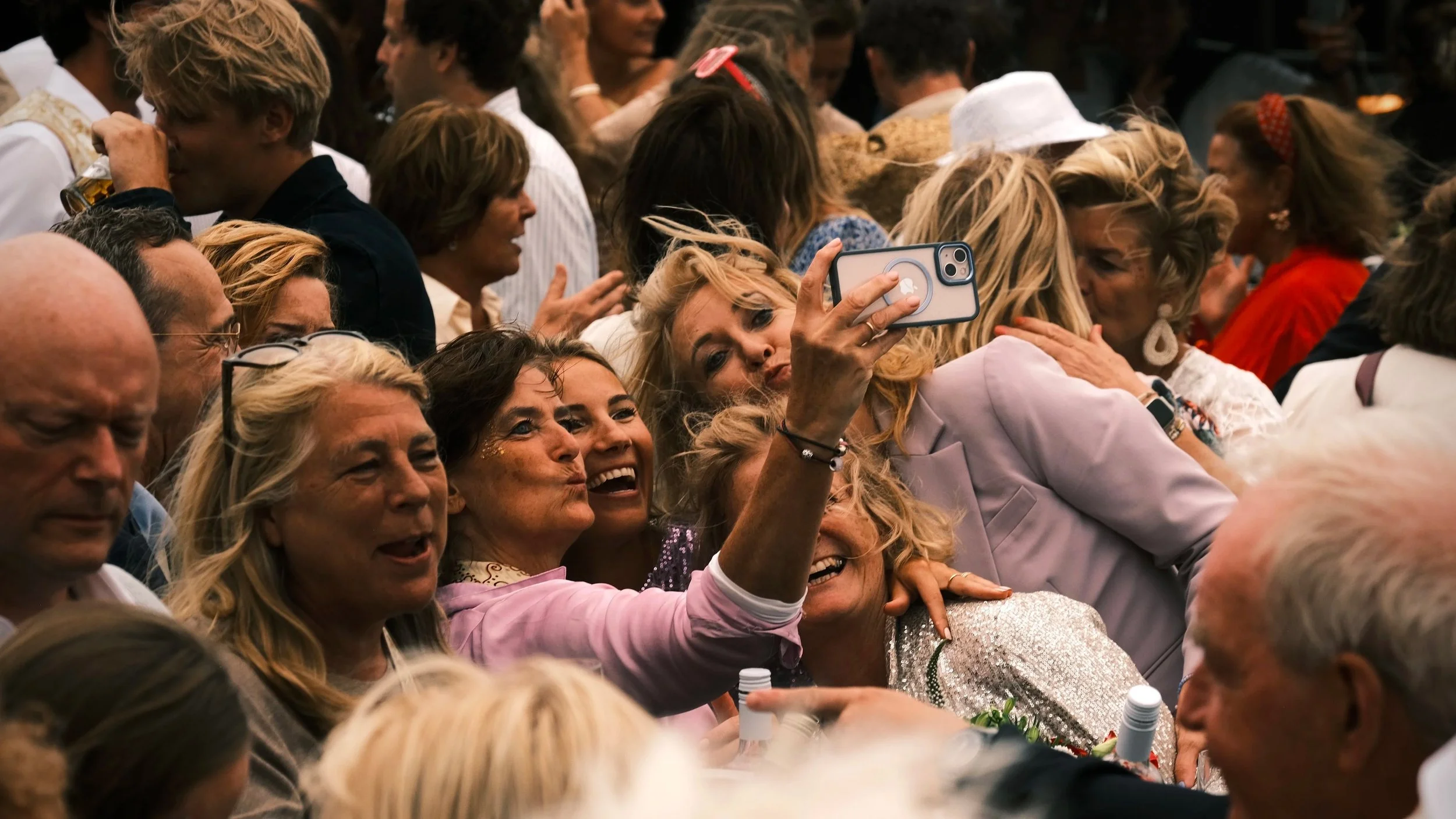 Een groep mensen zit dicht bij elkaar en lacht terwijl een vrouw met blond haar een smartphone gebruikt om een foto te maken.