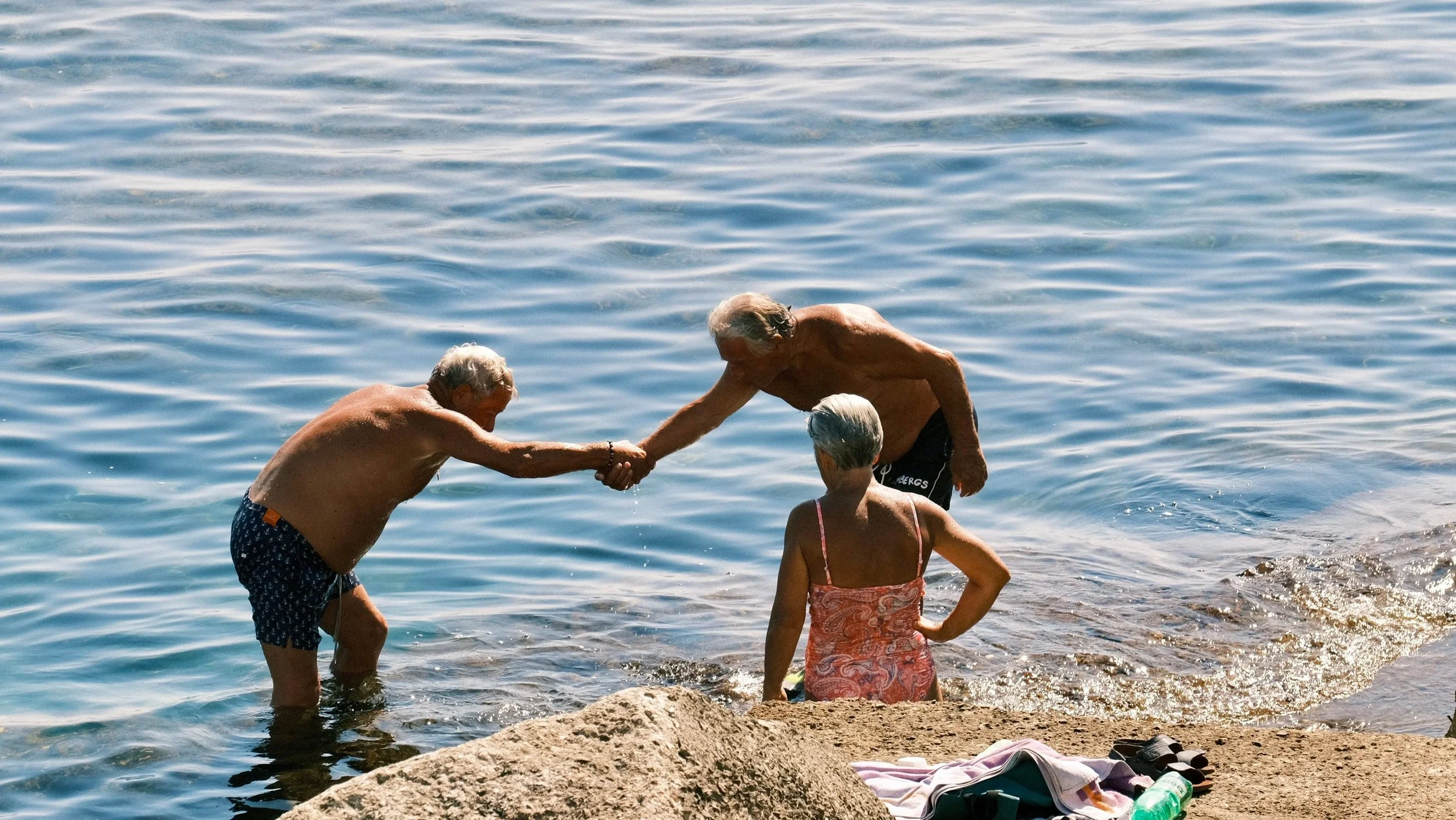 Twee ouderen helpen elkaar uit de zee tijdens een zonnige dag op het strand, met een vrouw die wacht op de rand van het water en spullen op de rots