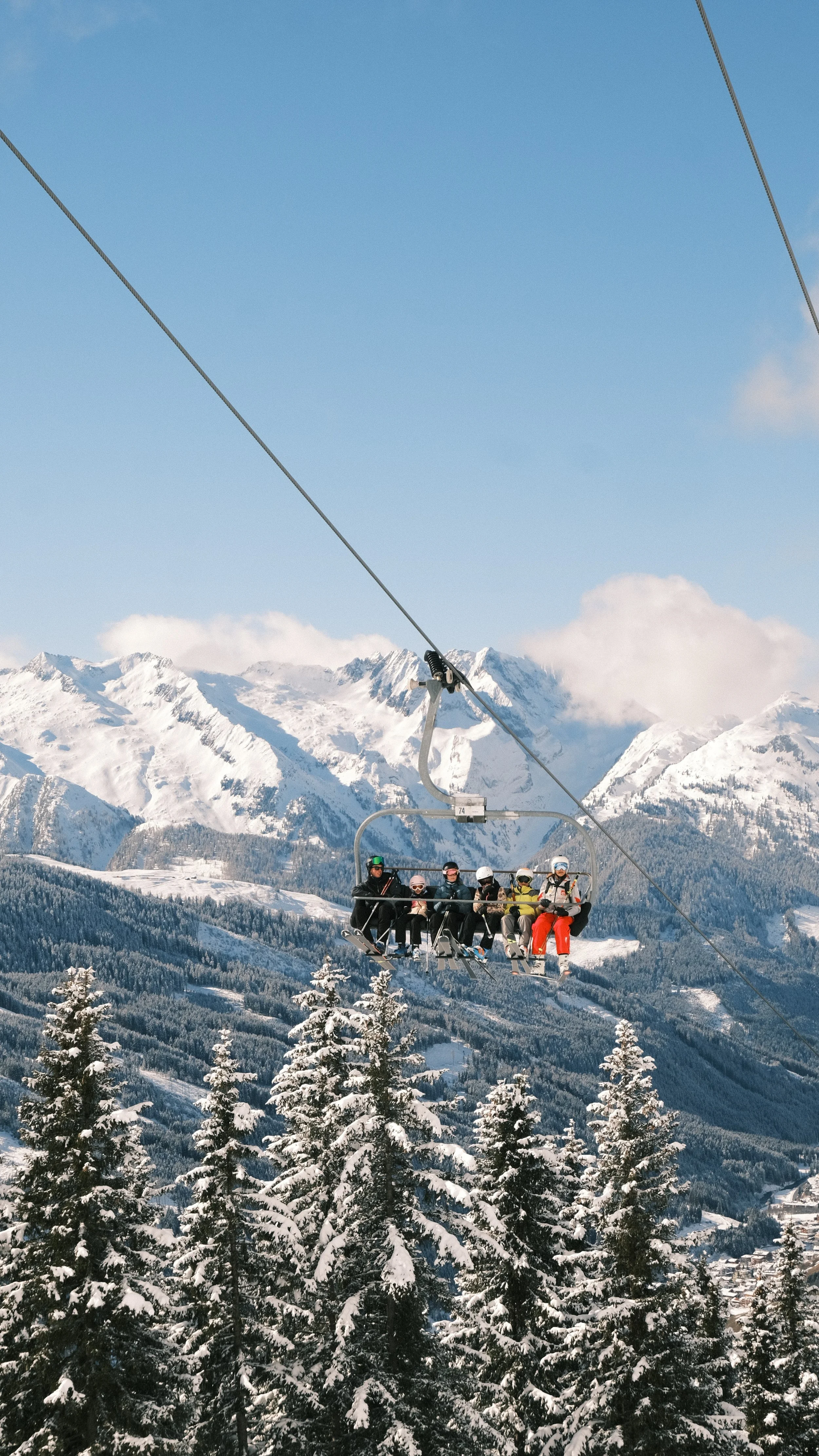A group of people riding a ski lift in a snow-covered mountain landscape with pine trees and snow-capped peaks in the background.