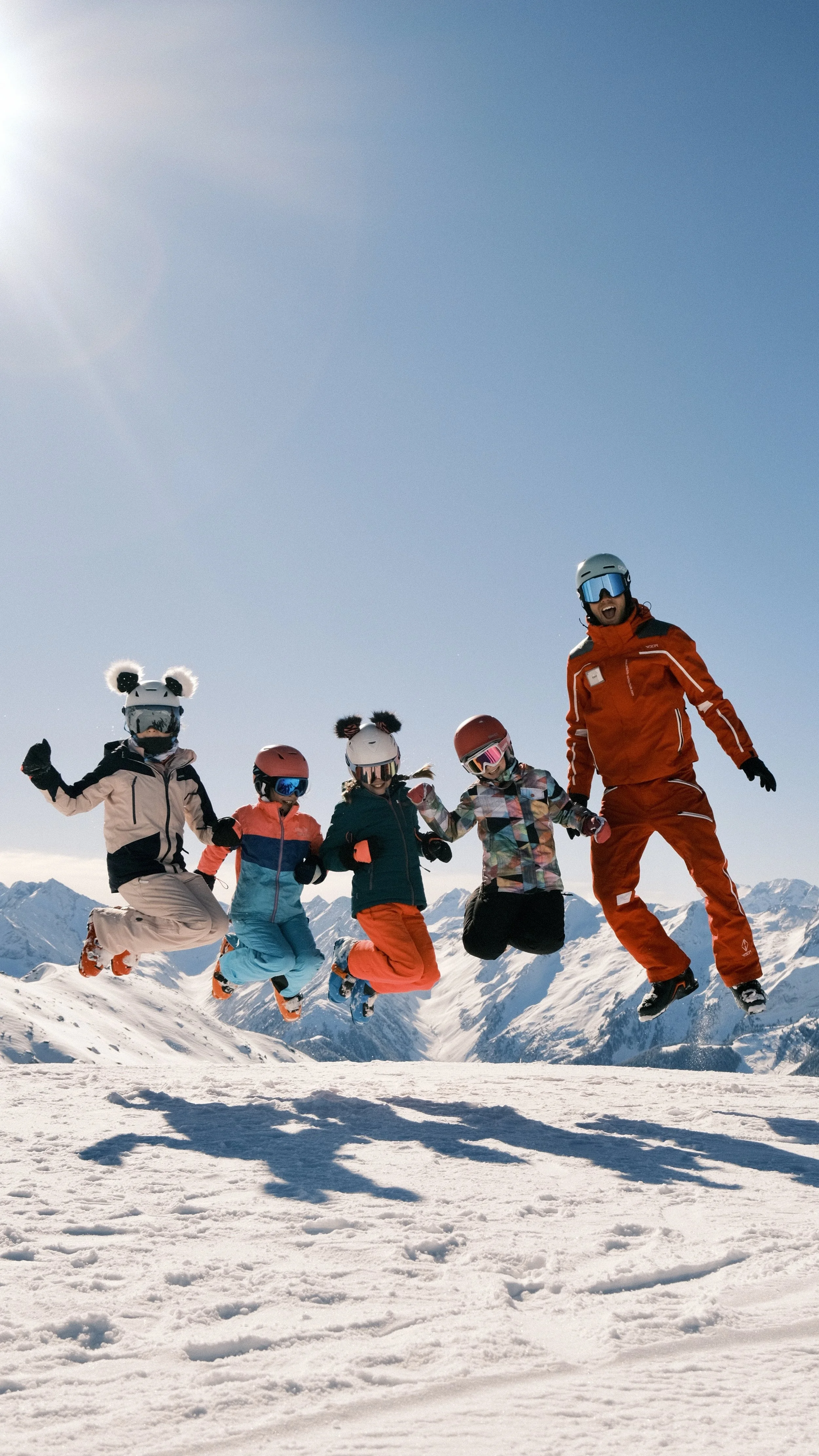 Five children and an adult jumping in the snow on a mountain range during a sunny day, all wearing snow gear and helmets.