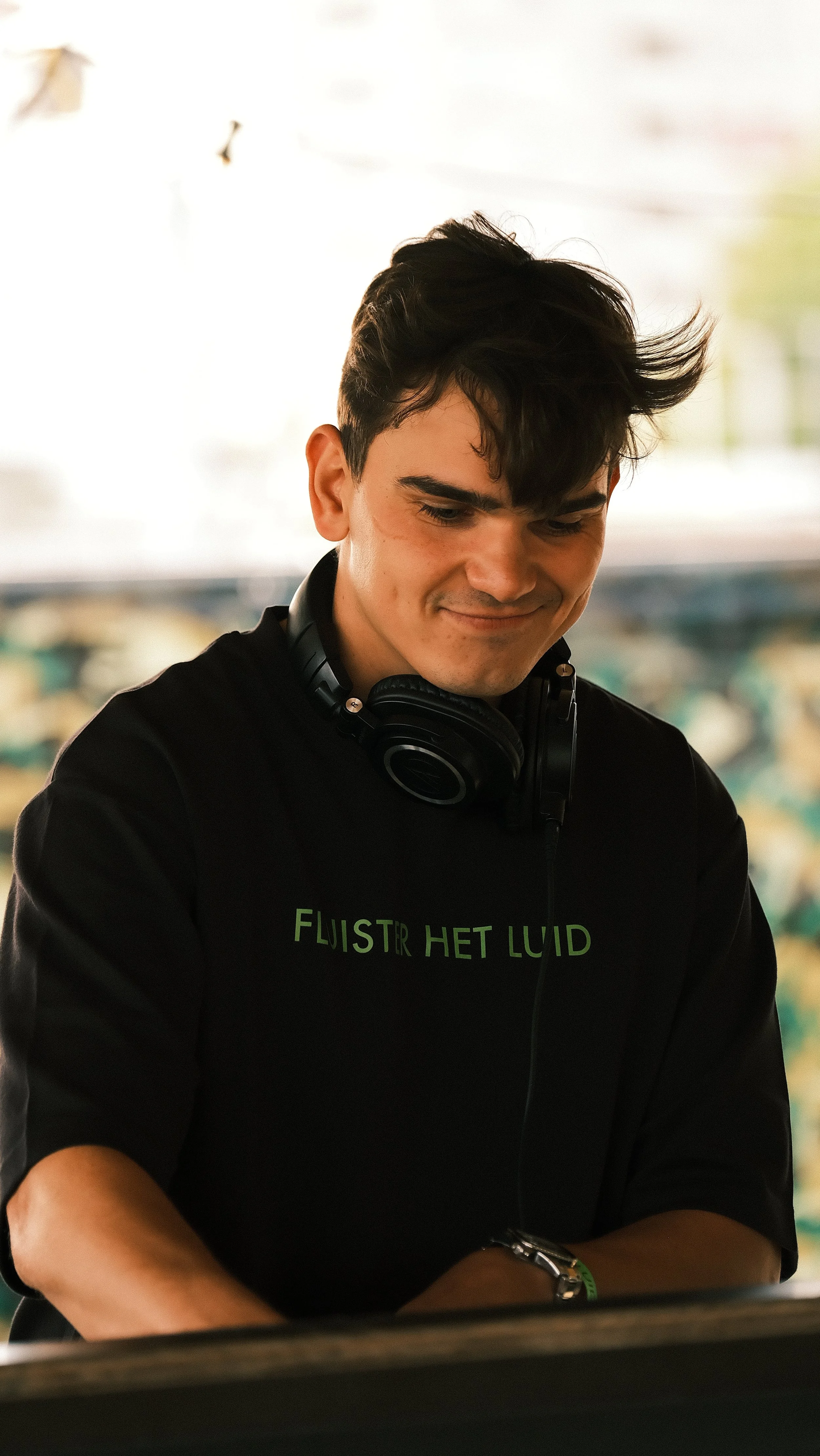Portret van een jonge man met zwart haar, lachend en er plezier in hebbend, draagend een zwarte hoodie met groene tekst en een koptelefoon om zijn nek.