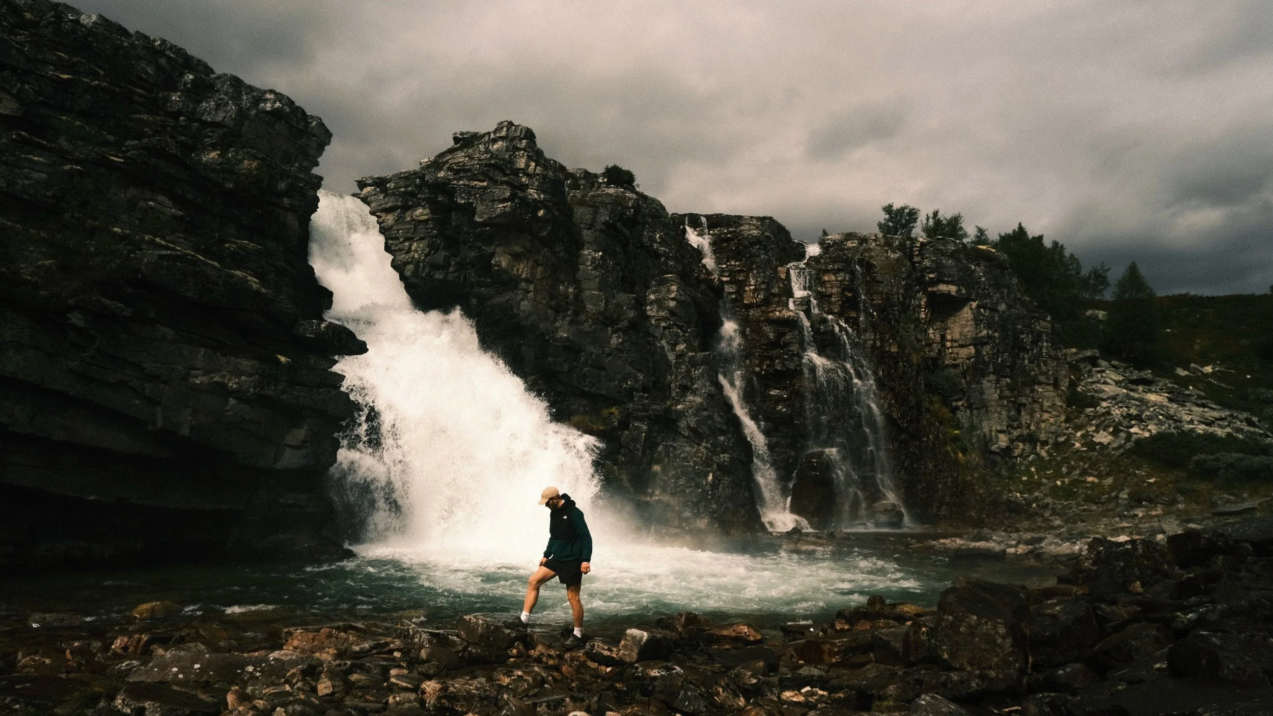 Person standing on rocks in front of a large waterfall with rocky cliffs and trees in the background on a cloudy day.