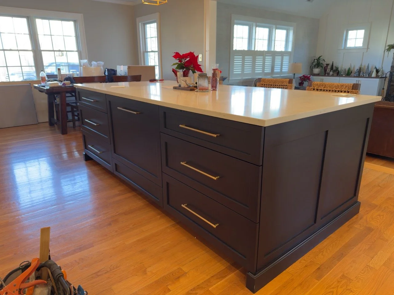 A kitchen island with a dark-colored base and a light-colored countertop, set in a bright room with hardwood floors and large windows. Decorative items and plants are on a side shelf and table nearby.