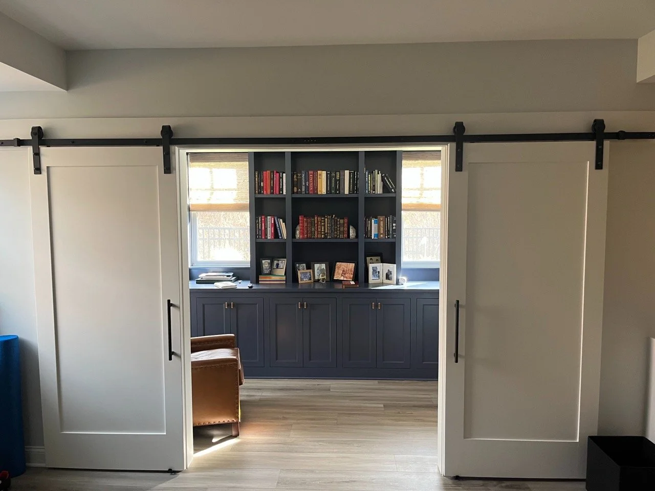 View of a room through sliding barn doors, showing a dark blue bookshelf with books and framed photos, flanked by two windows with blinds, and a chair partially visible in the foreground.