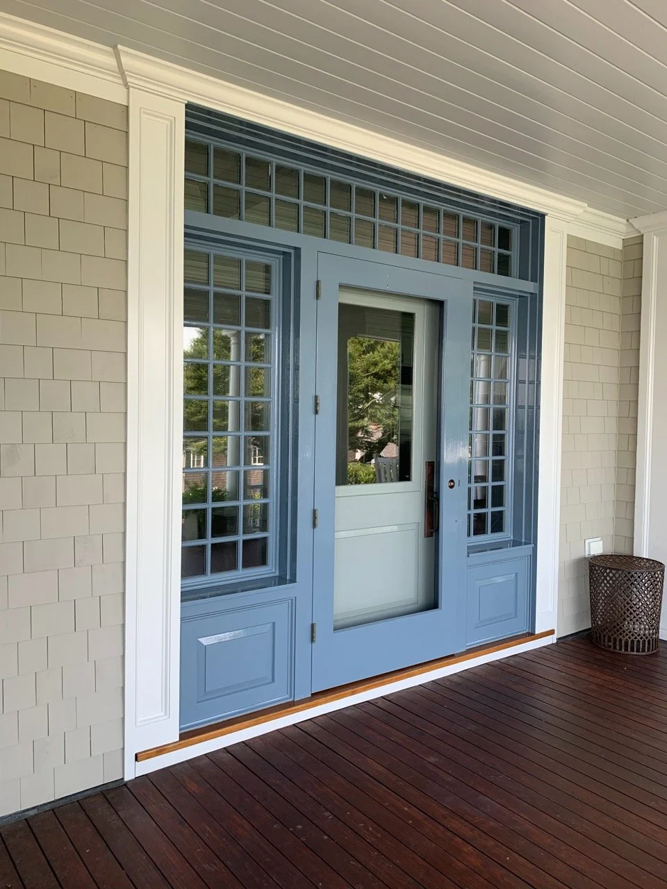 Blue front door with glass panels, surrounded by beige siding and white trim, on a porch with dark wooden flooring.