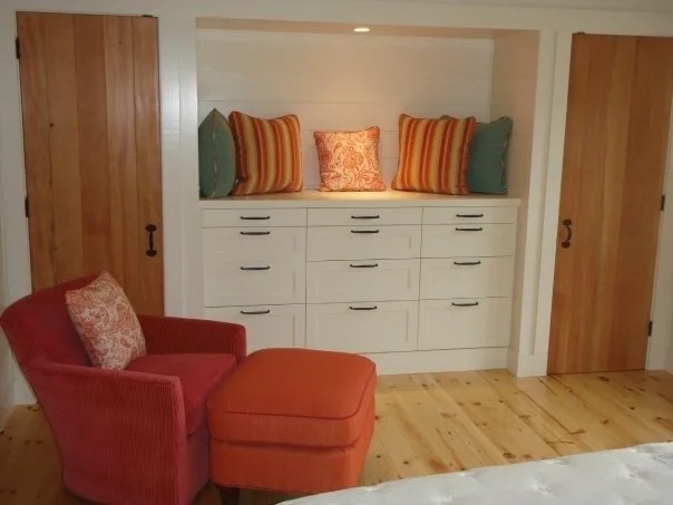 A built-in white dresser with multiple drawers, topped with four decorative pillows in orange and green, built into a recessed wall in a room with wooden doors and flooring. An orange armchair with a patterned pillow and matching ottoman is in front 