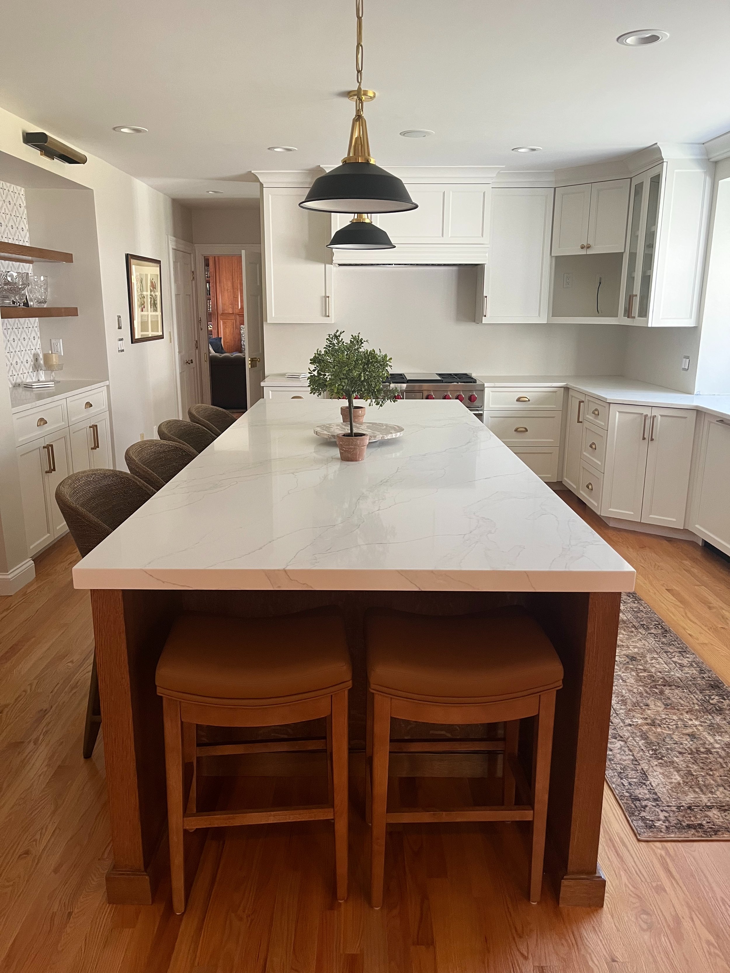A modern kitchen with a white marble island, black and gold pendant lights, white cabinets, and a potted plant on the island.