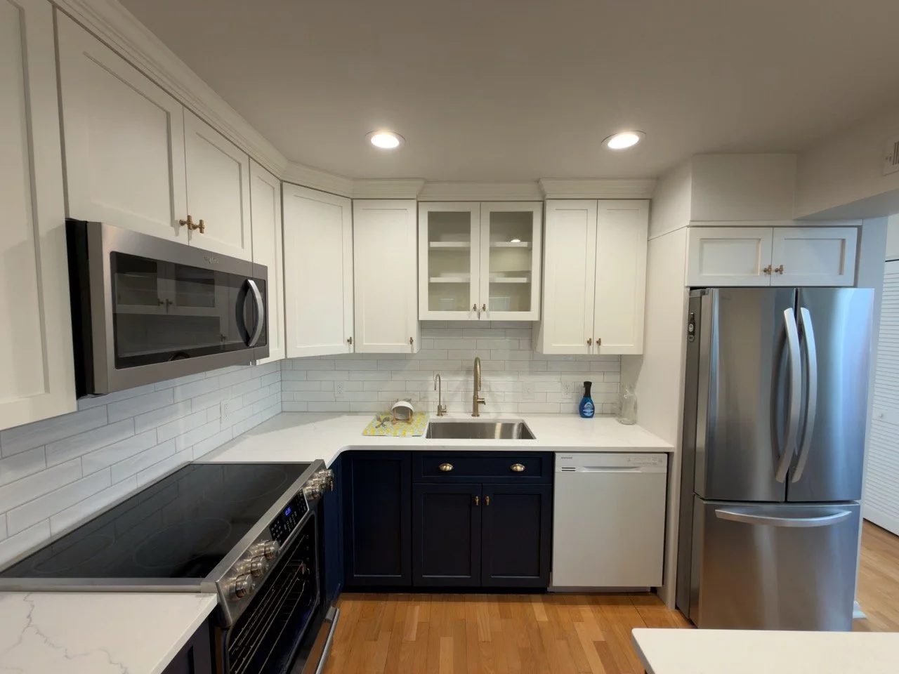 Kitchen with white and navy cabinets, stainless steel microwave, refrigerator, and oven, white countertops, and white subway tile backsplash, hardwood floors, and ceiling recessed lighting.