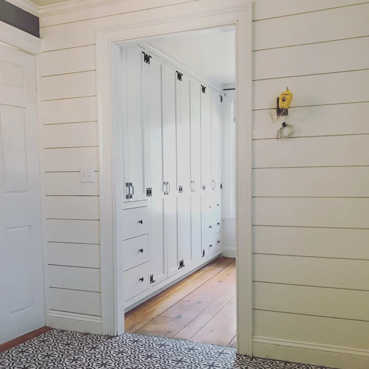 A hallway with white shiplap walls, a patterned black-and-white floor, and a white built-in wardrobe with black hardware in the background.